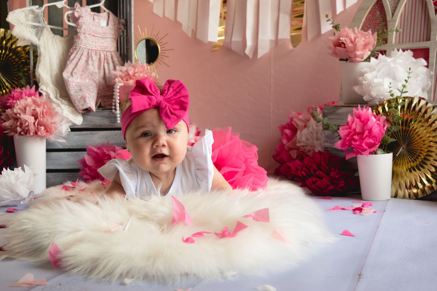 Baby wearing pink bow lies on fluffy white rug with pink decorations and gold accents in background.