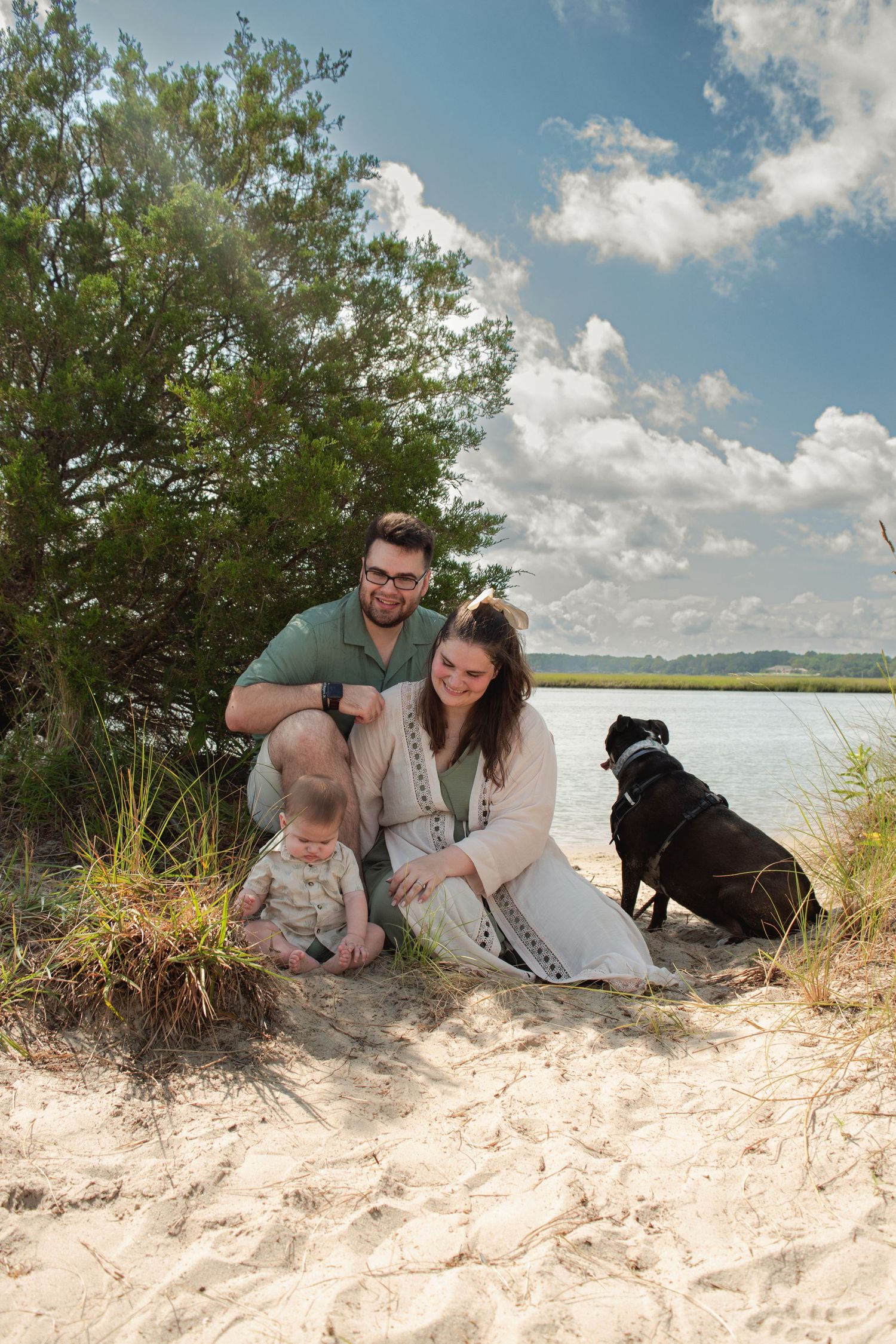Family with baby and black dog sitting on sandy beach near water and trees under partly cloudy sky.