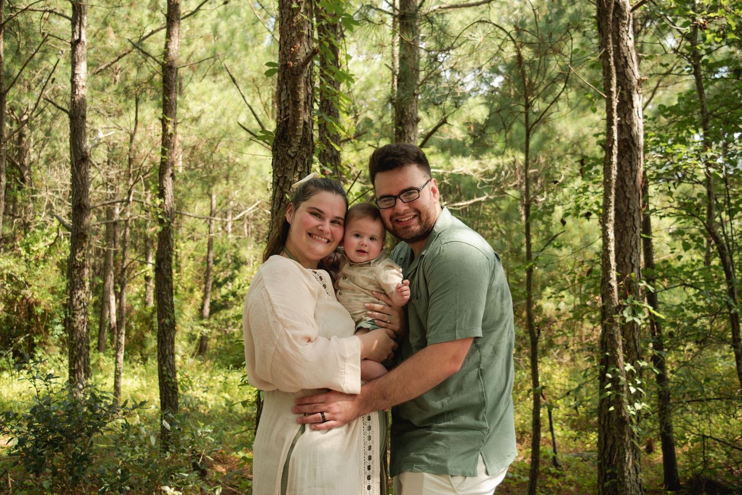 Young family with baby poses smiling in lush green forest setting for outdoor portrait photography.