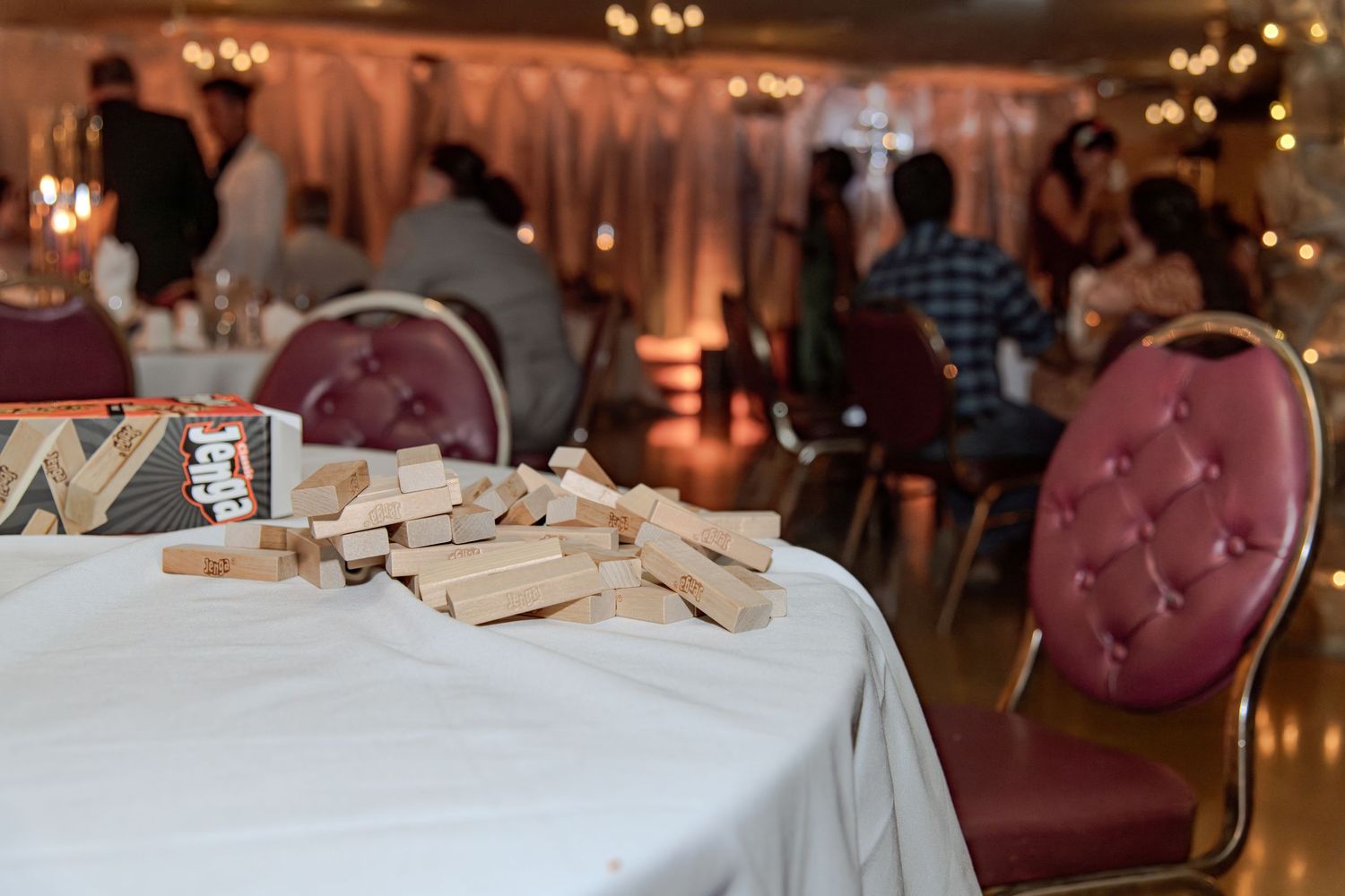 Jenga blocks scattered on a white tablecloth at an elegant wedding reception with guests and fairy lights.