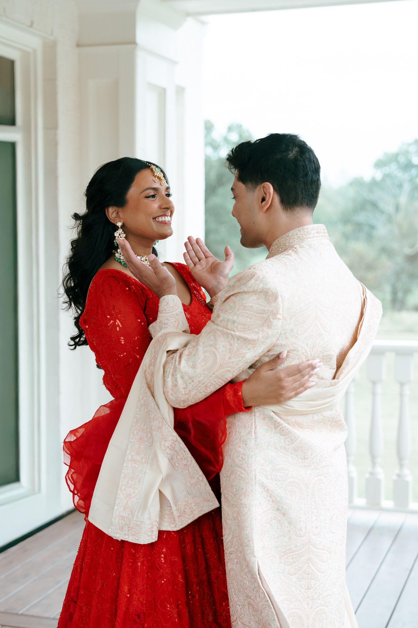 A couple in traditional South Asian wedding attire share a joyful moment on a white porch, with the bride in red and groom in cream.