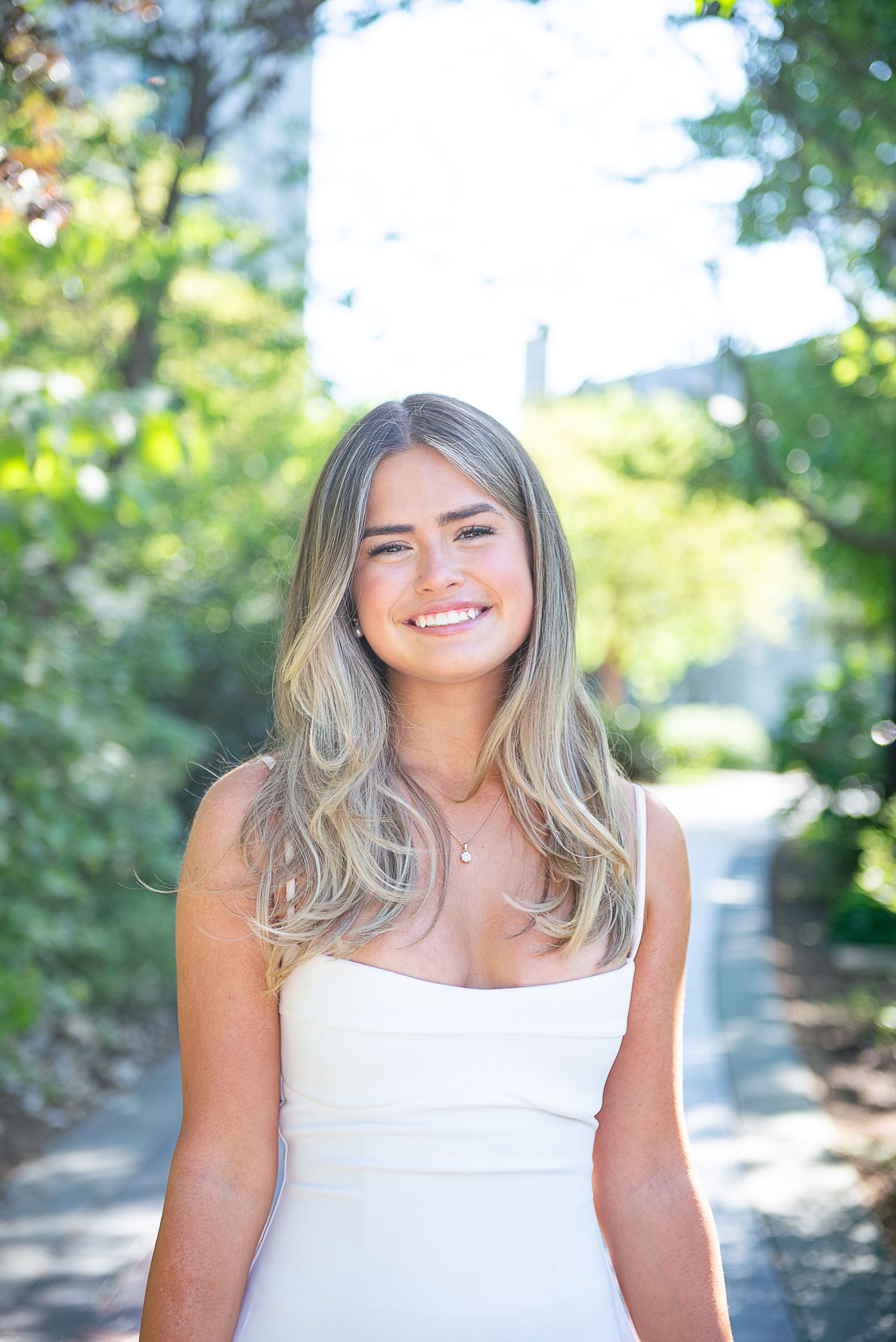 Graduate in white dress poses for portrait with sunlit trees and greenery in background on Villanova's campus.