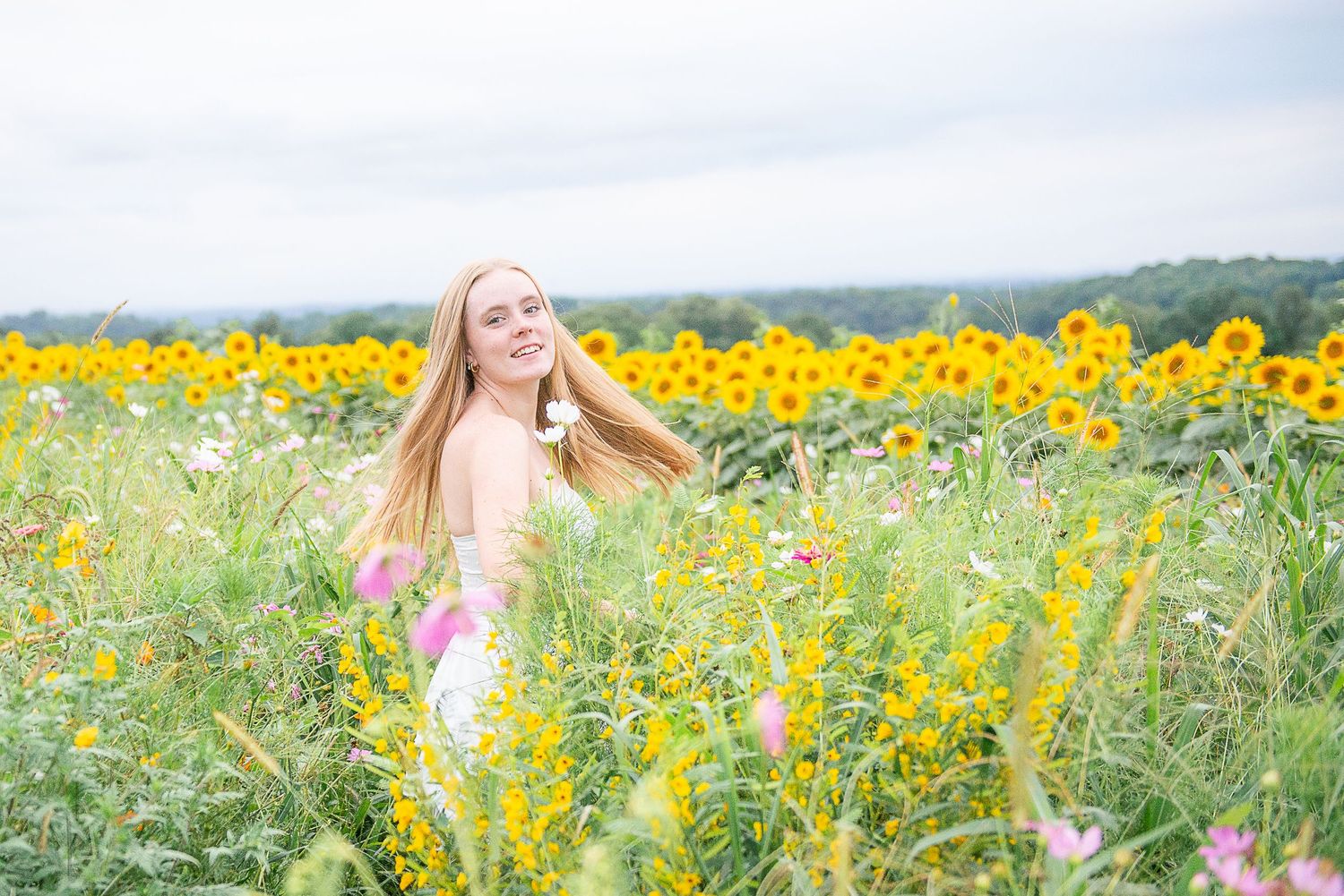 Young blonde woman smiling in a vibrant sunflower and wildflower field on a cloudy day.
