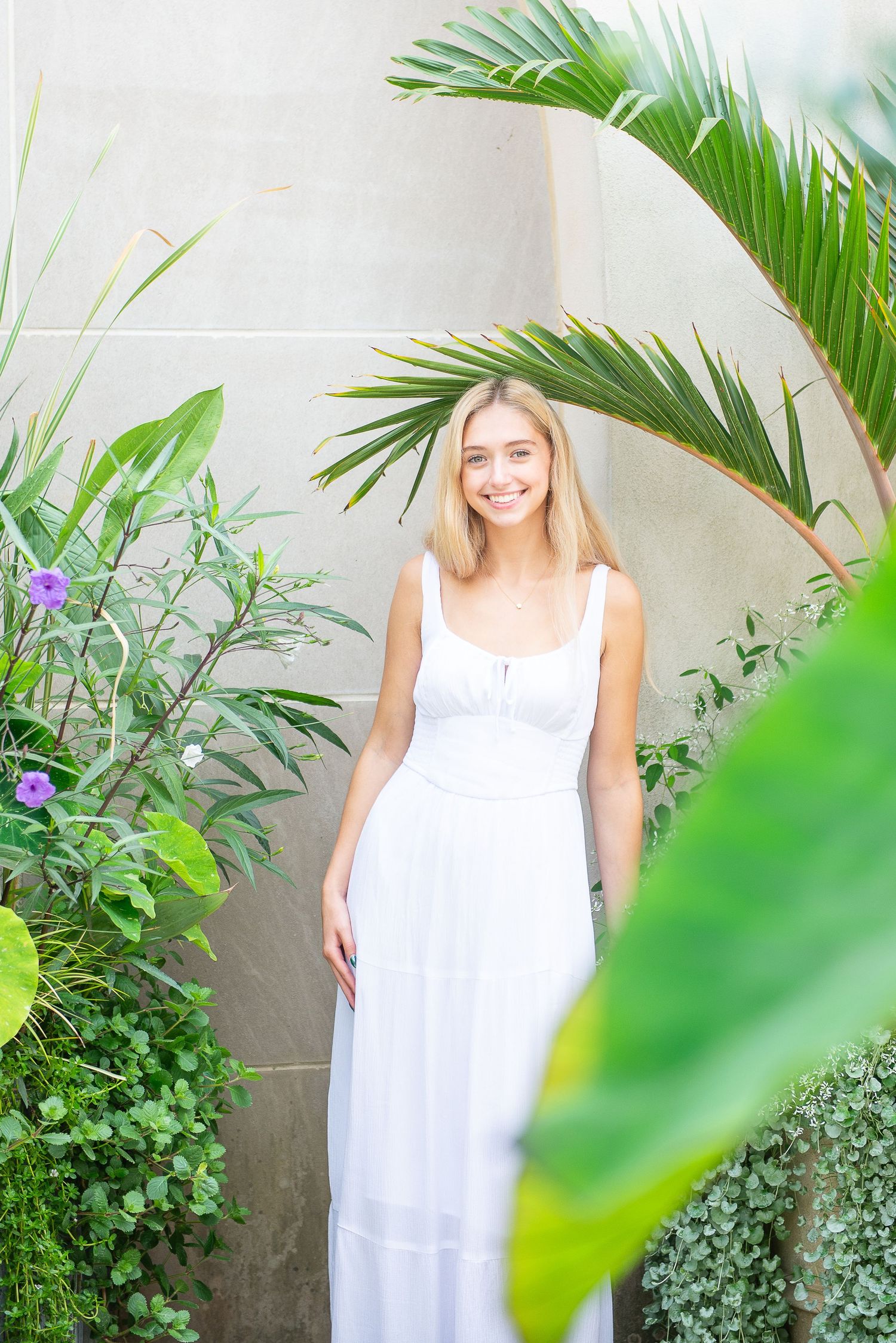 A flowing white maxi dress stands out against lush tropical plants and palm fronds in an outdoor garden setting.