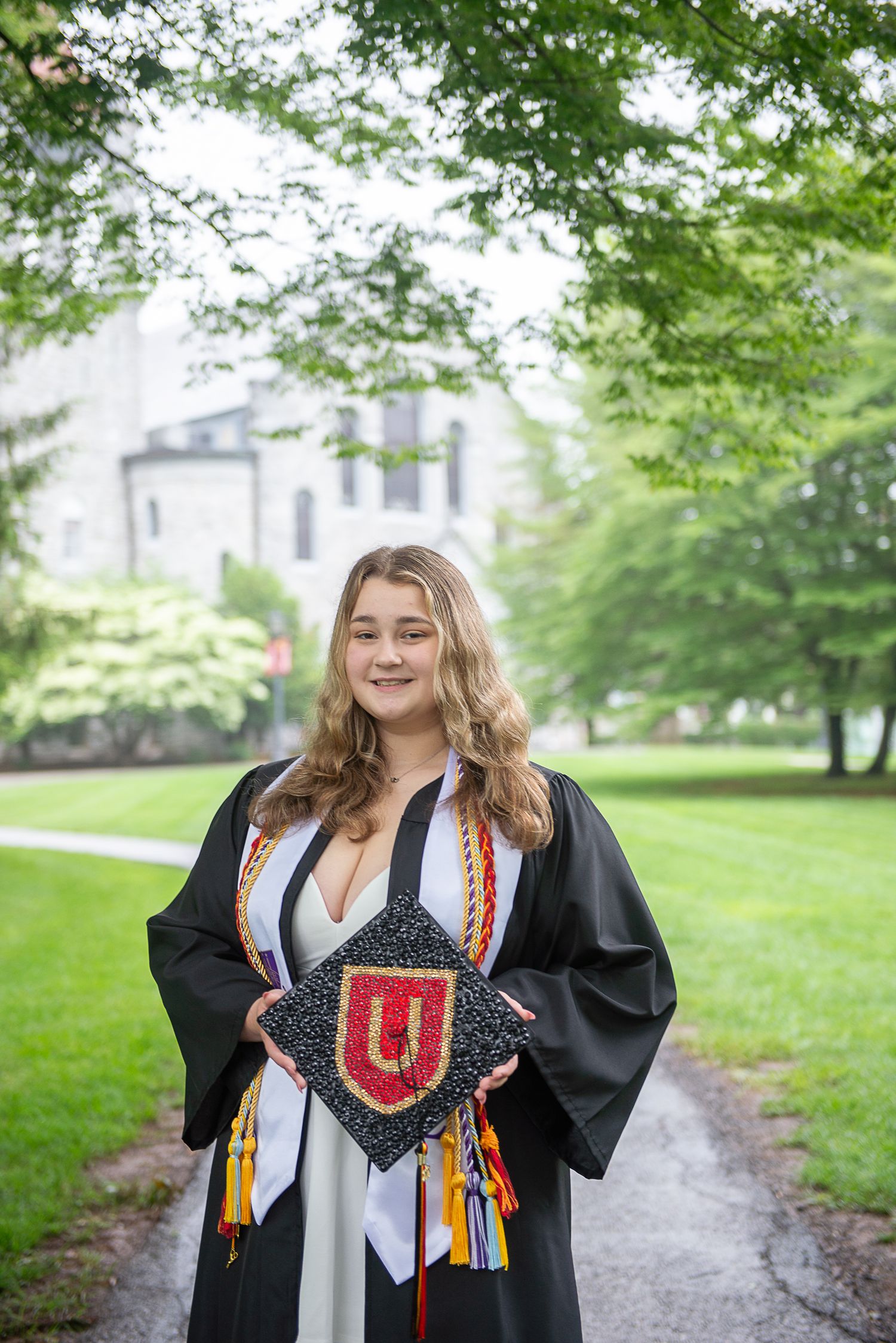 Graduate in black cap and gown holding decorated graduation cap with red Ursinus College logo on campus lawn.