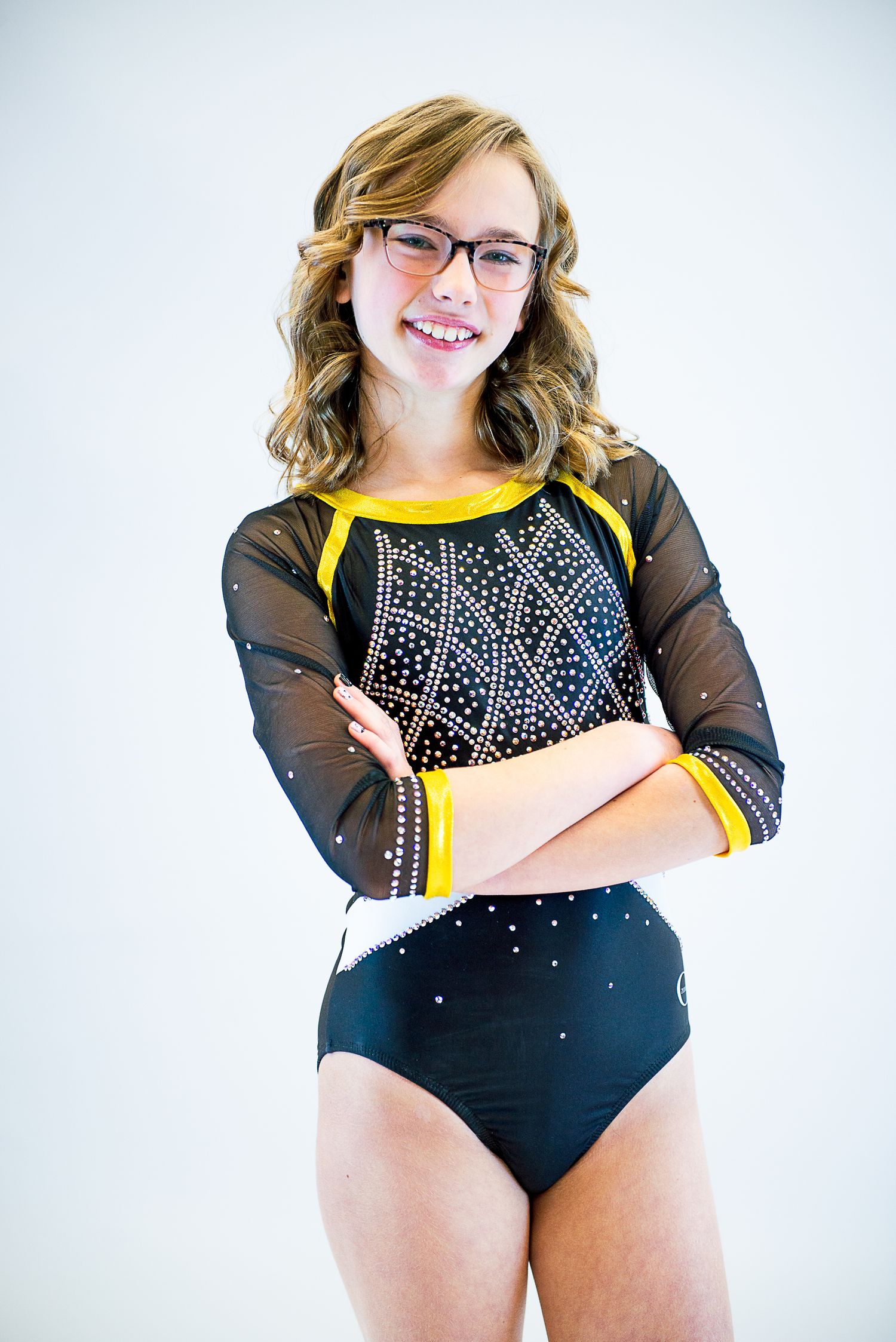 Gymnast in black sparkly leotard with mesh sleeves poses against white background.