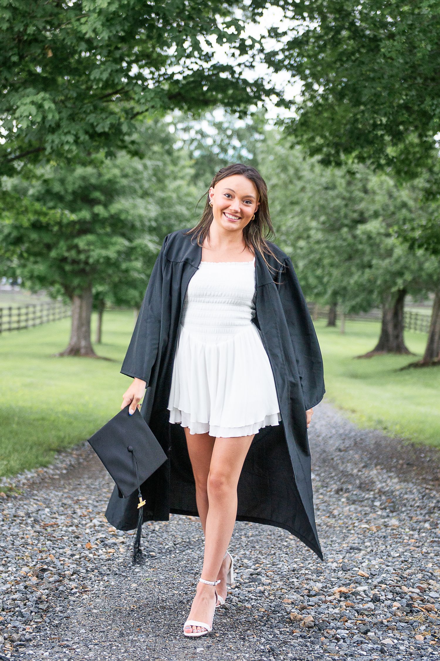 Graduate in white dress and black graduation gown poses on gravel path surrounded by trees.