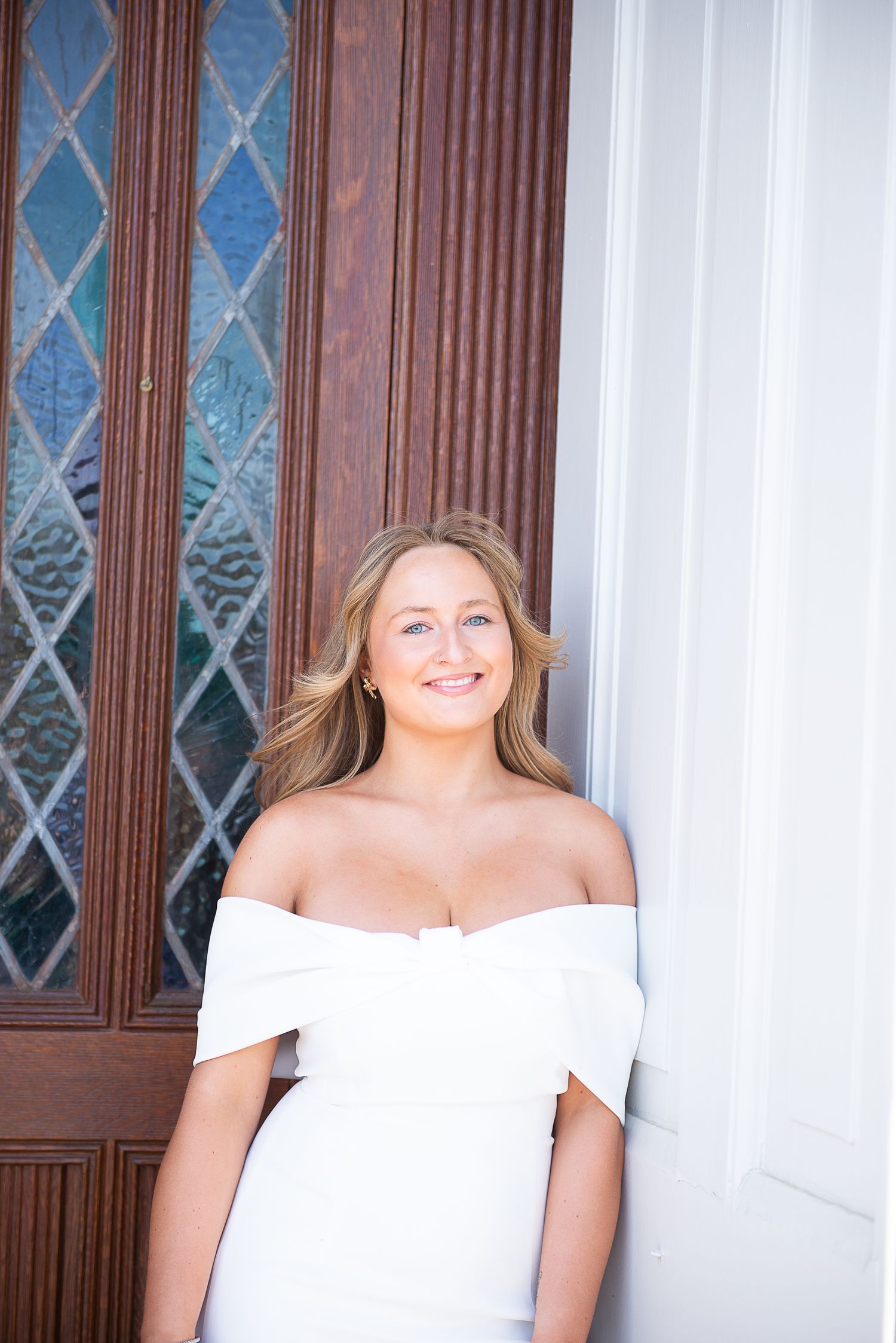 Graduate in elegant white off-shoulder dress poses by ornate wooden door.