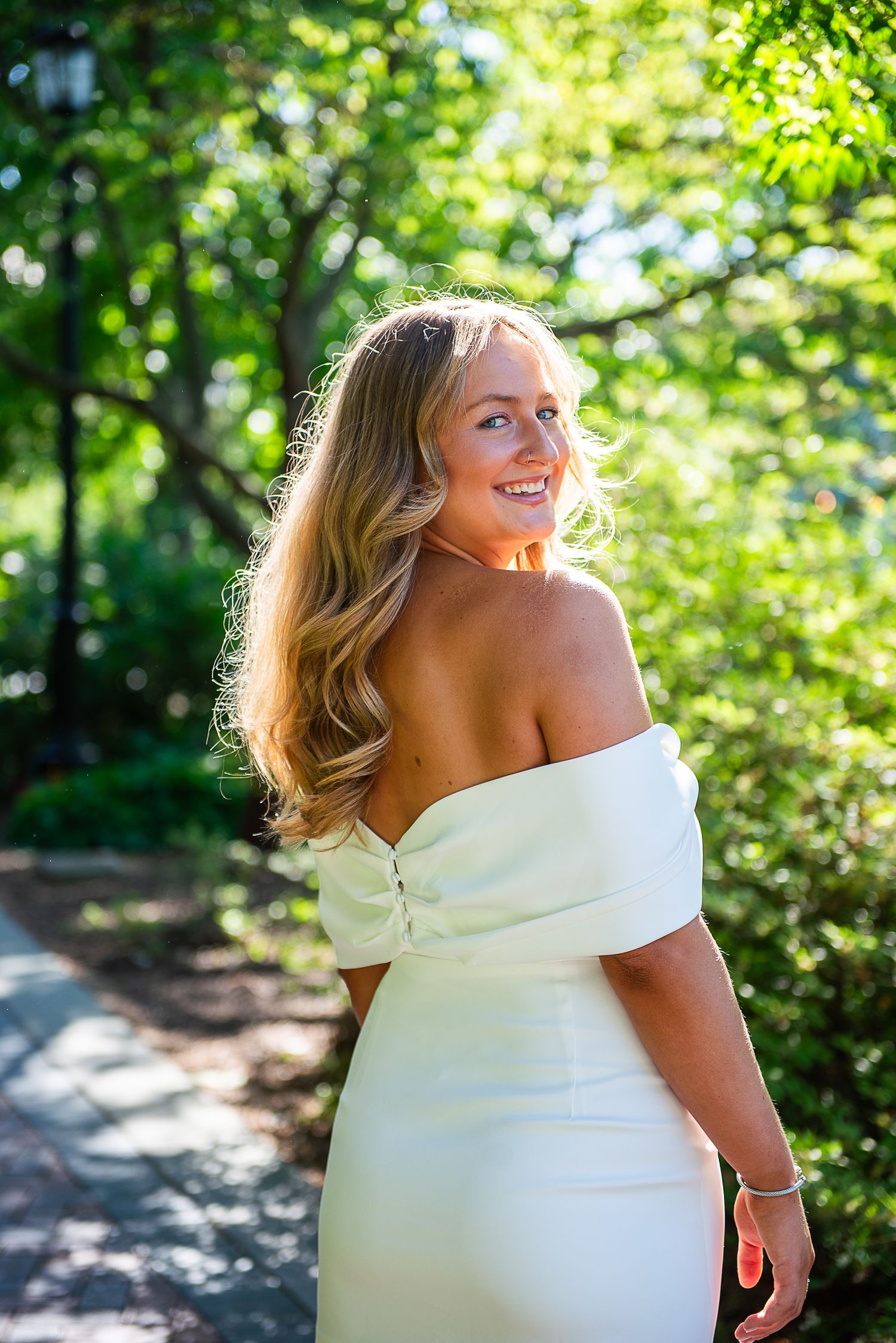 Graduate in elegant white dress poses outdoors with sunlit greenery in background creating a dreamy effect.
