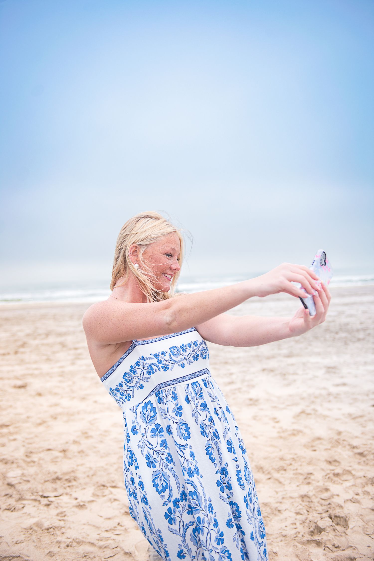 Blonde woman in blue floral sundress taking a selfie on a sandy beach with ocean in background.