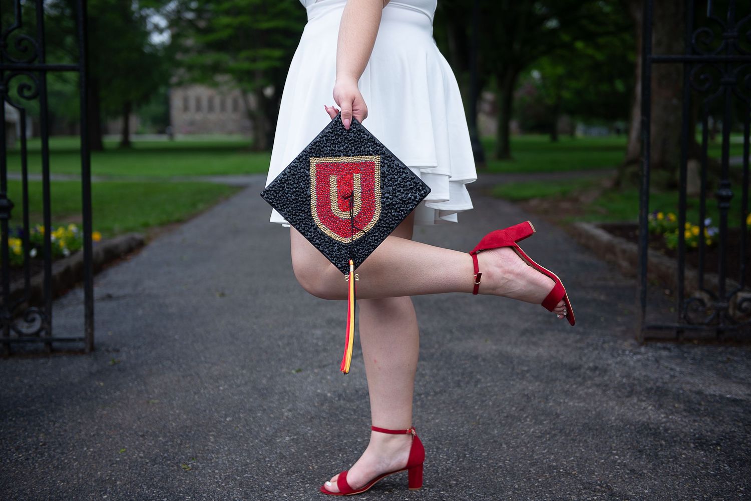 Graduate lifts leg showing red heels while holding decorated graduation cap with Ursinus College logo.