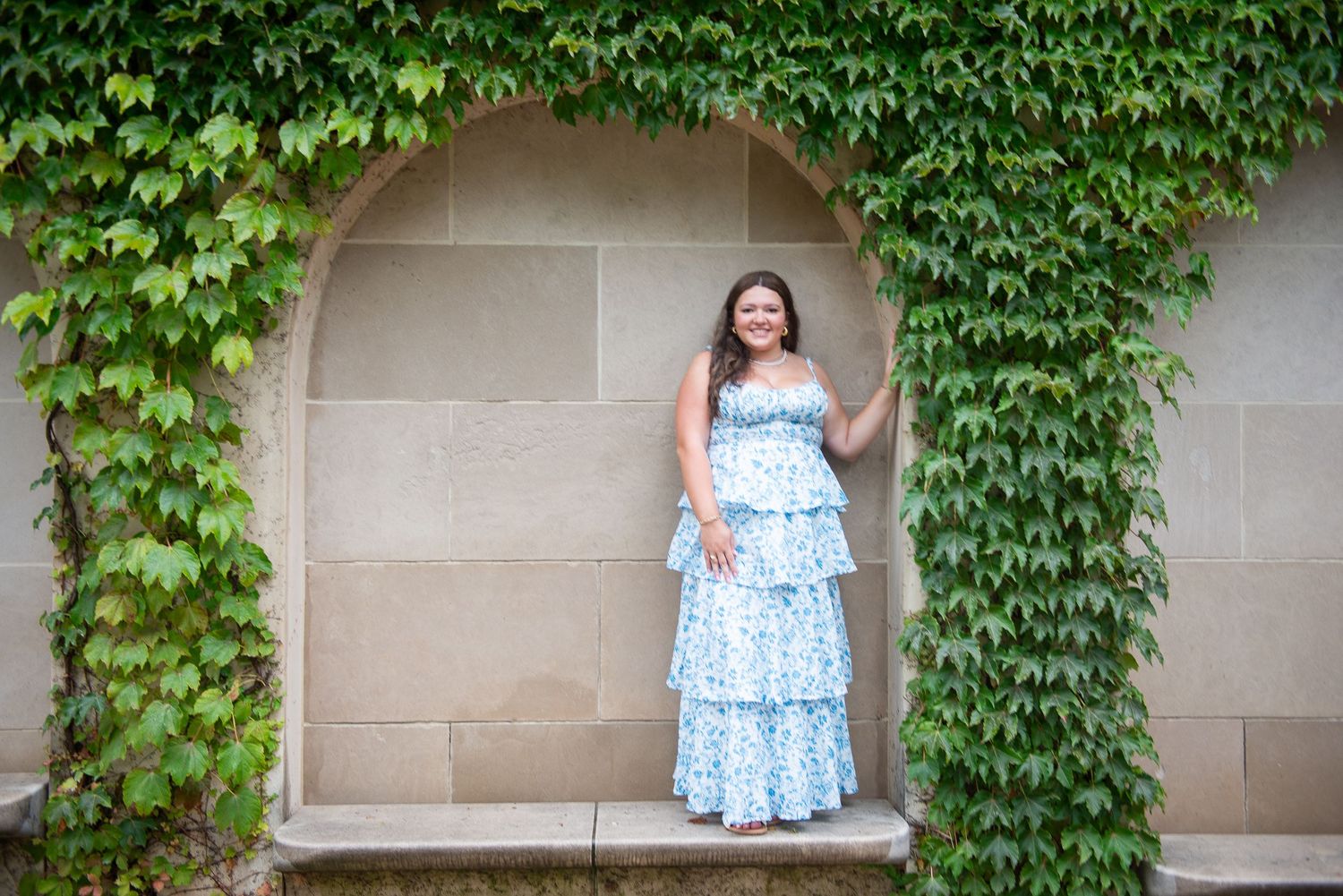 A tiered blue dress creates contrast against ivy-covered stone walls in an alcove setting.