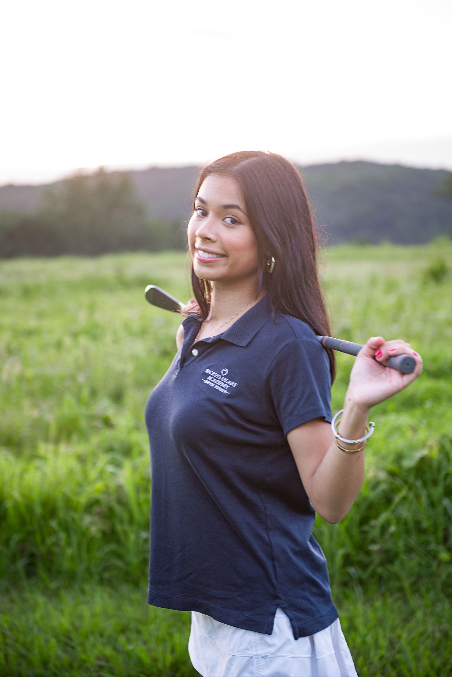 A navy polo-clad golfer poses with a club over their shoulder on a verdant course at dusk.