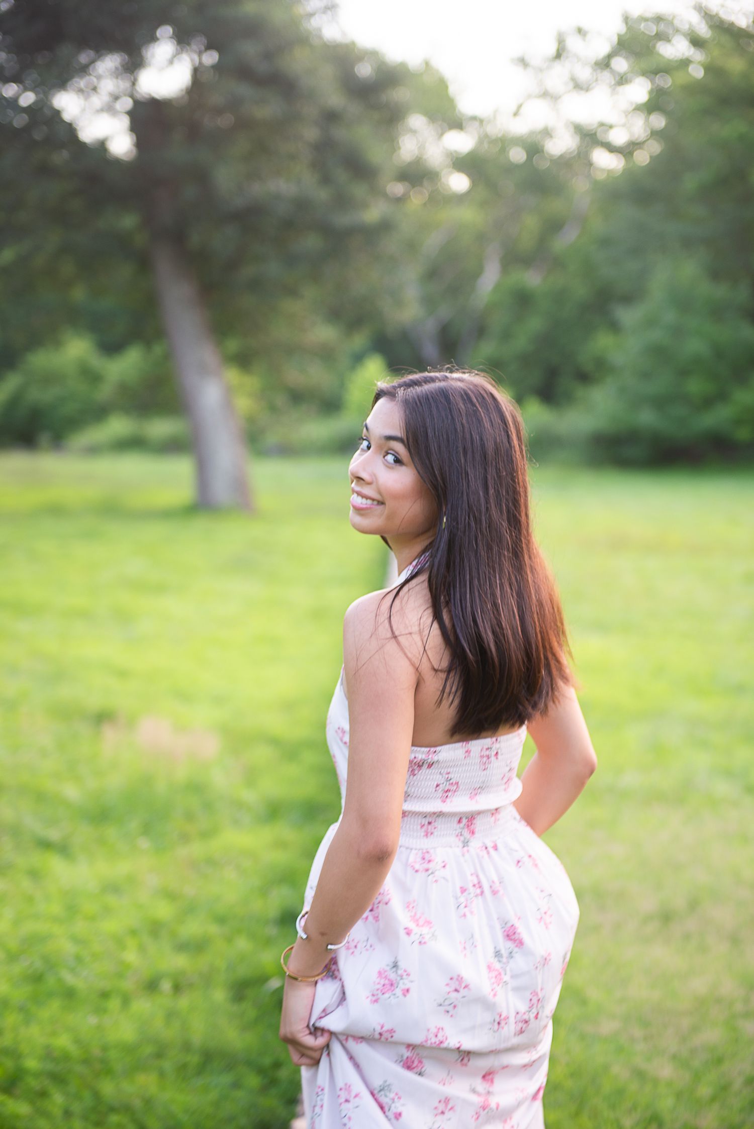 A senior in a floral dress catches the evening light in a serene garden setting with trees in the background.