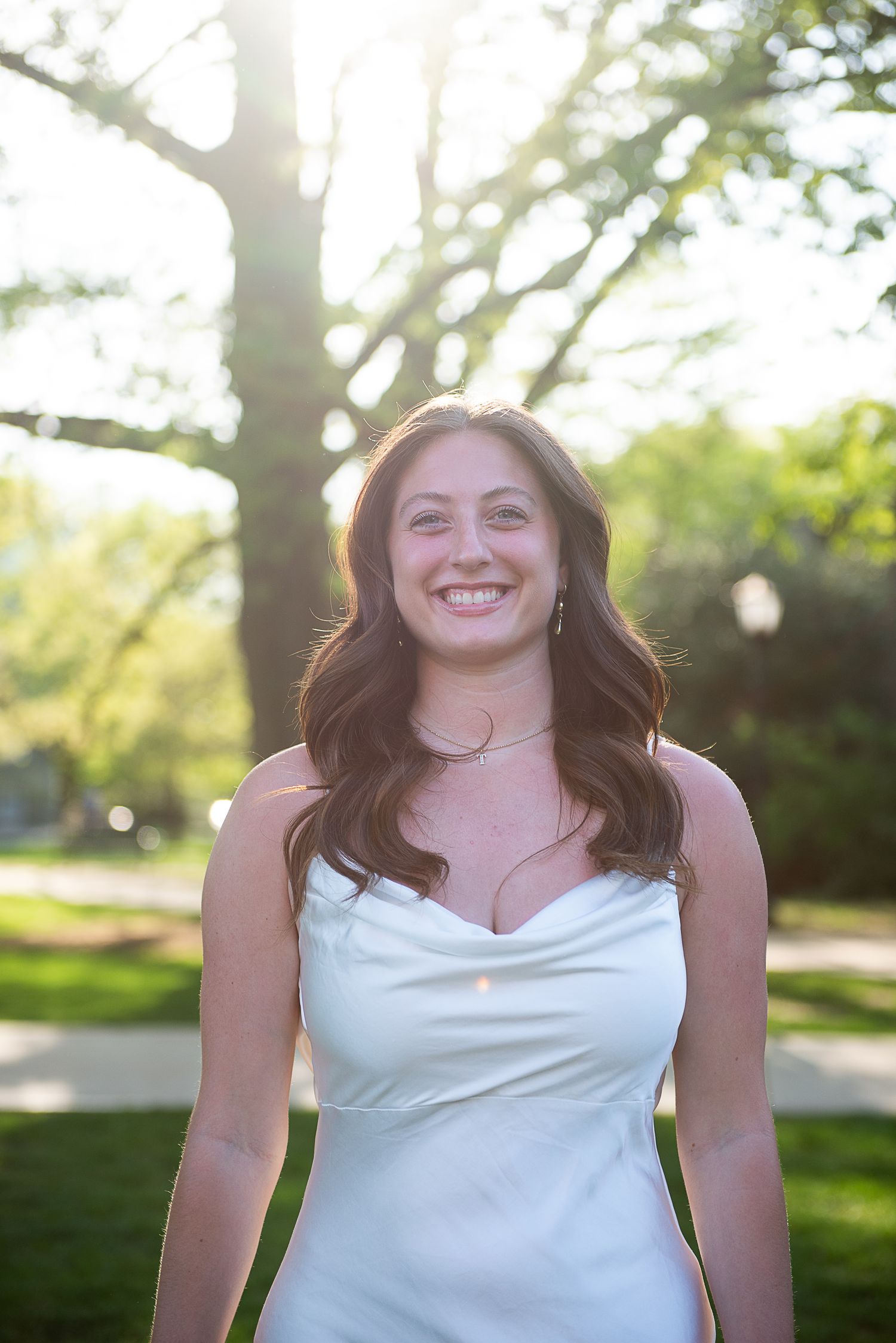 Graduation portrait in white dress captured outdoors with sunlit trees in background.