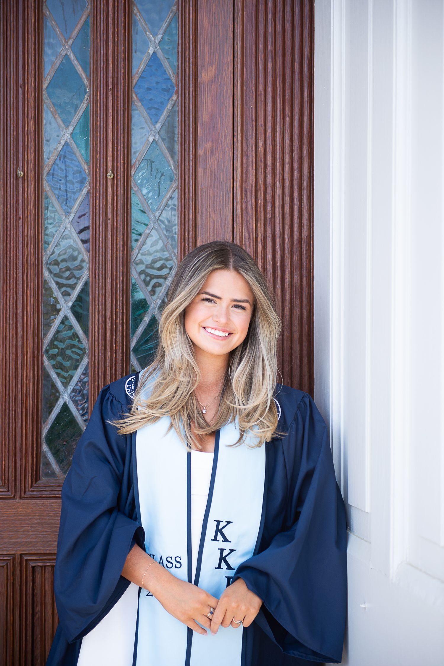Graduate in navy blue cap and gown poses in front of ornate wooden doors.