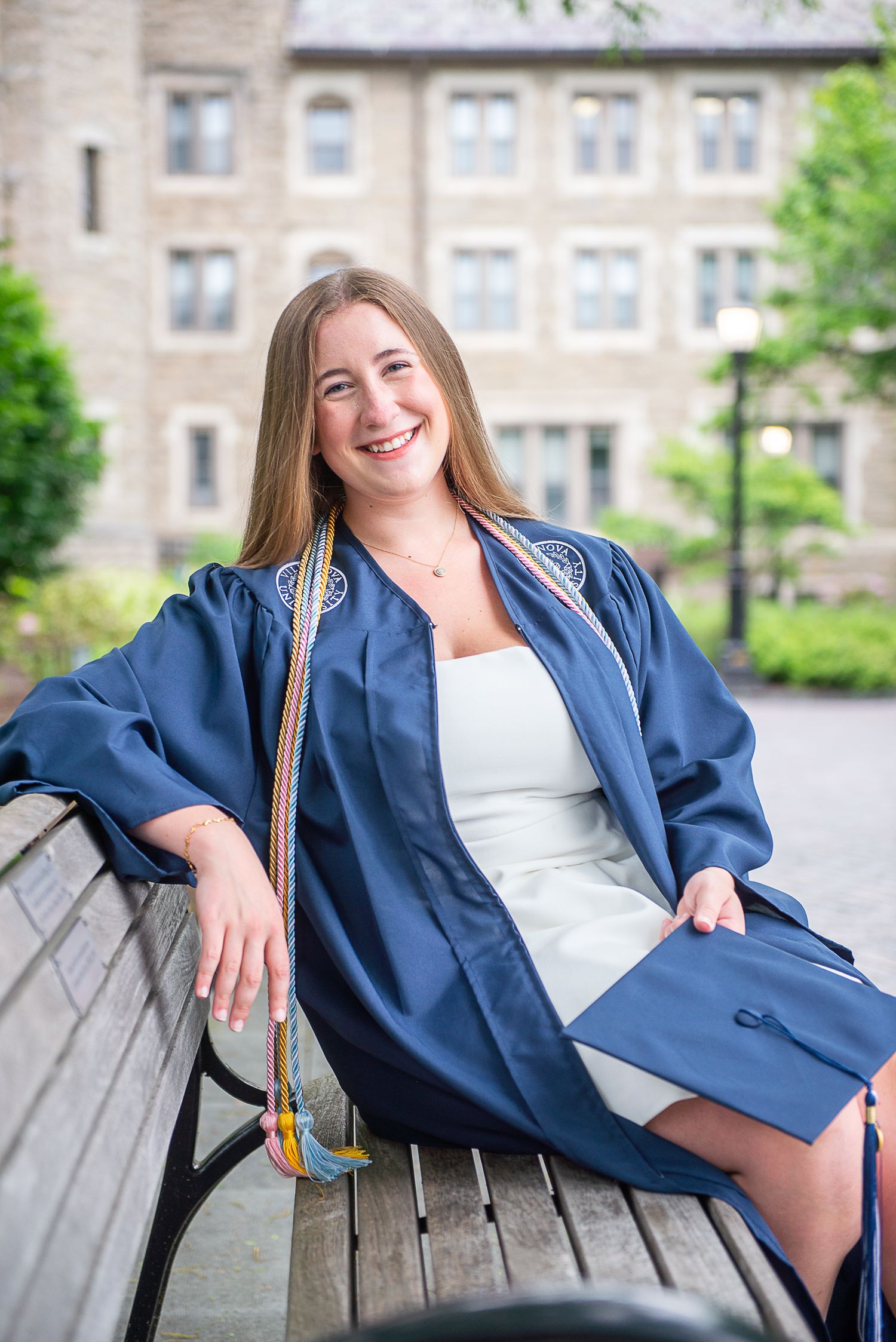 Graduate in blue gown sits casually on wooden bench with university building in background.