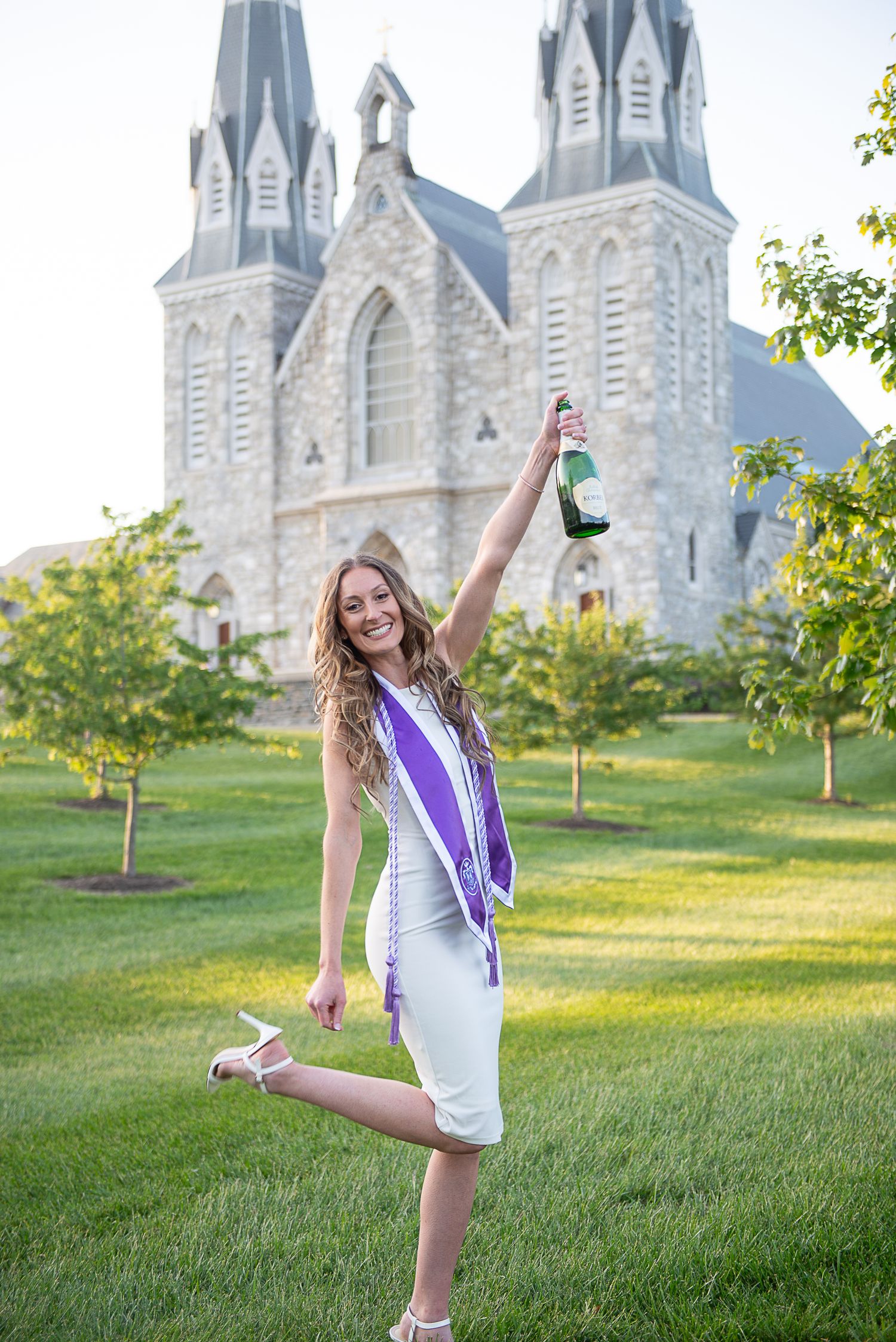 Graduate in white dress celebrates with champagne in front of St Thomas Church on Villanova's campus.
