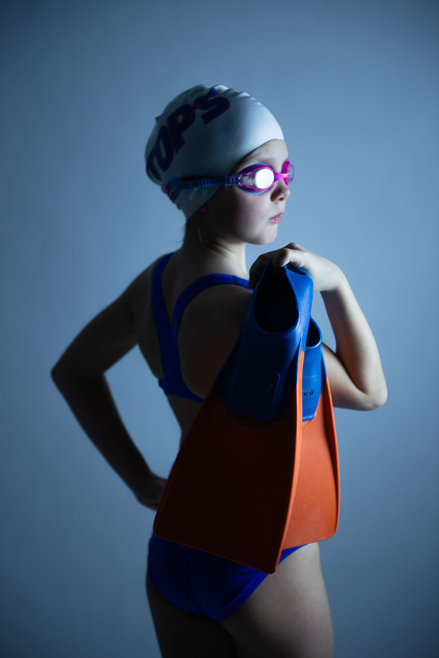 Dramatic portrait of swimmer in blue swimsuit and goggles against moody blue background.