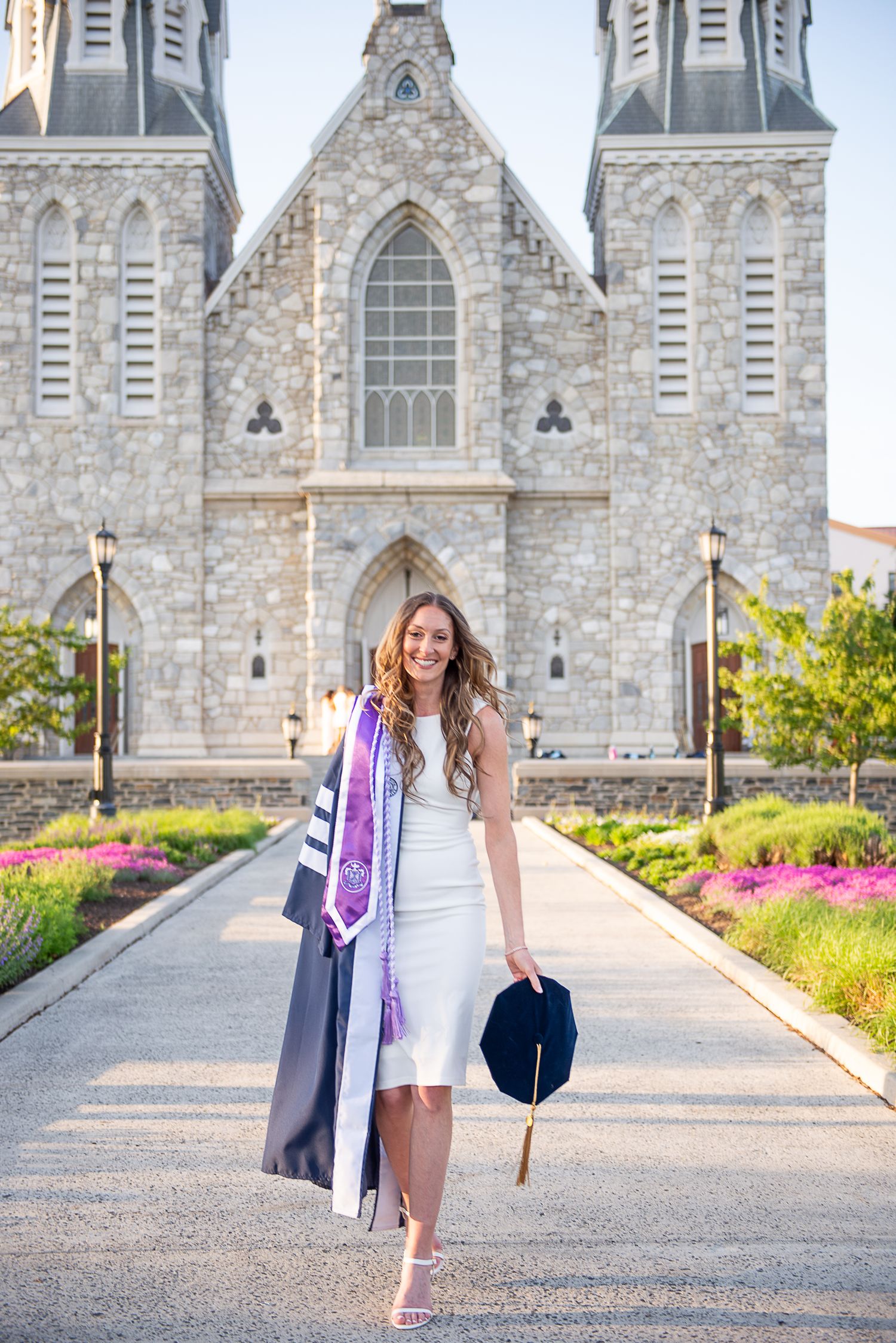 Graduate in white dress and graduation gown poses in front of St Thomas church at Villanova University with flowers in foreground.