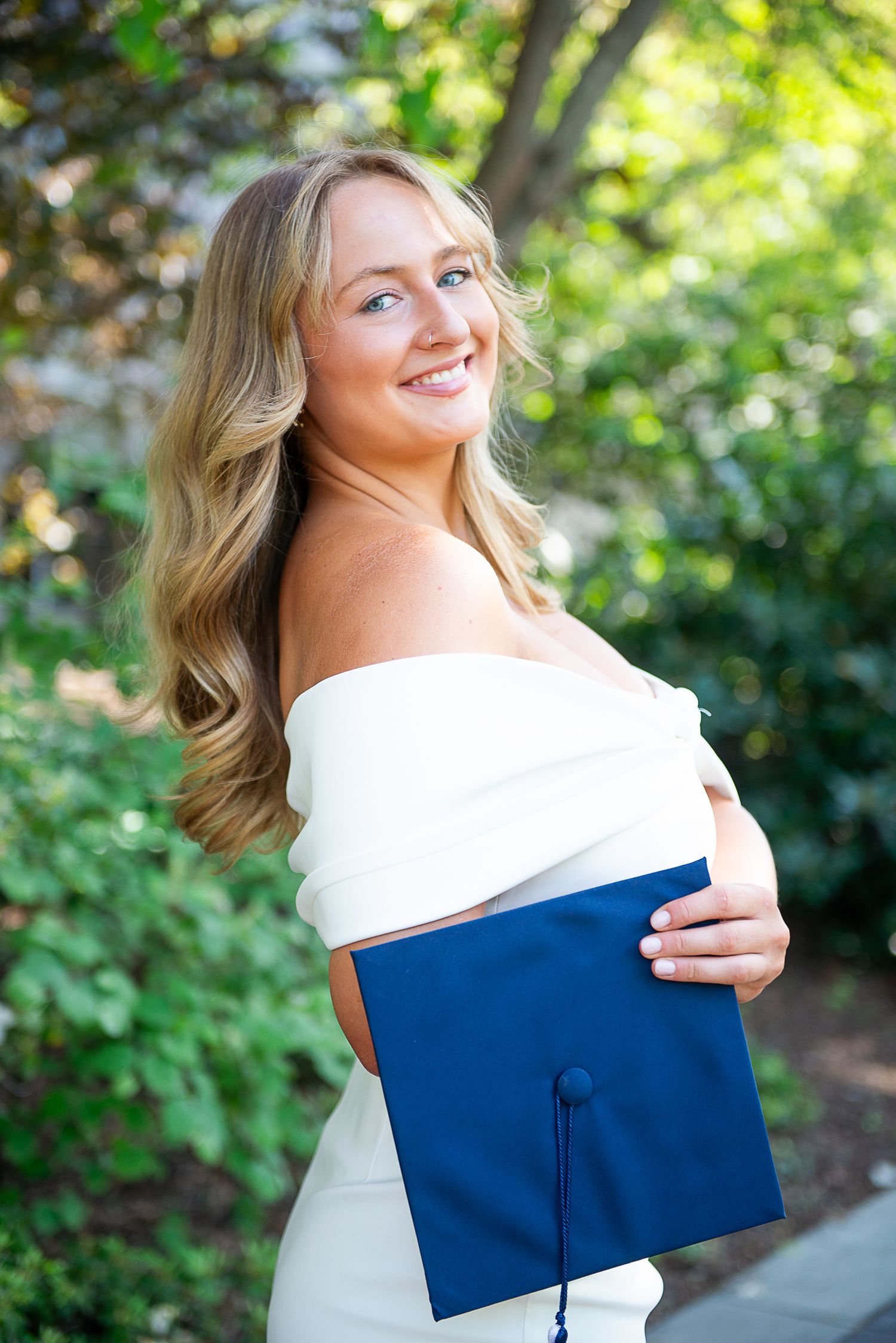 Graduate in white dress holds blue mortarboard cap against garden background.
