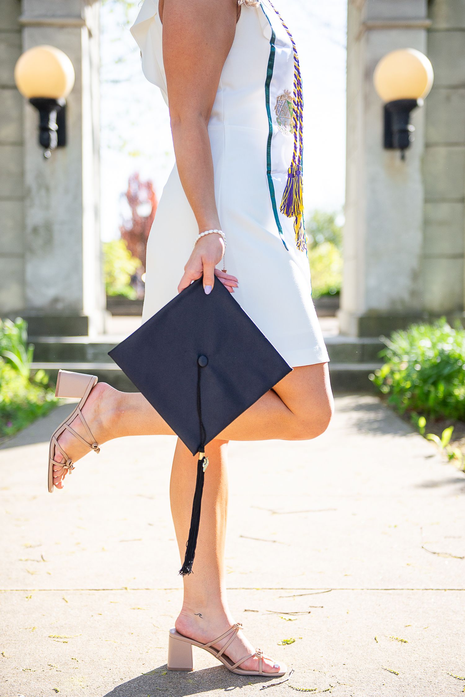 Graduate in white dress poses holding graduation cap while standing on one leg against stone archway.