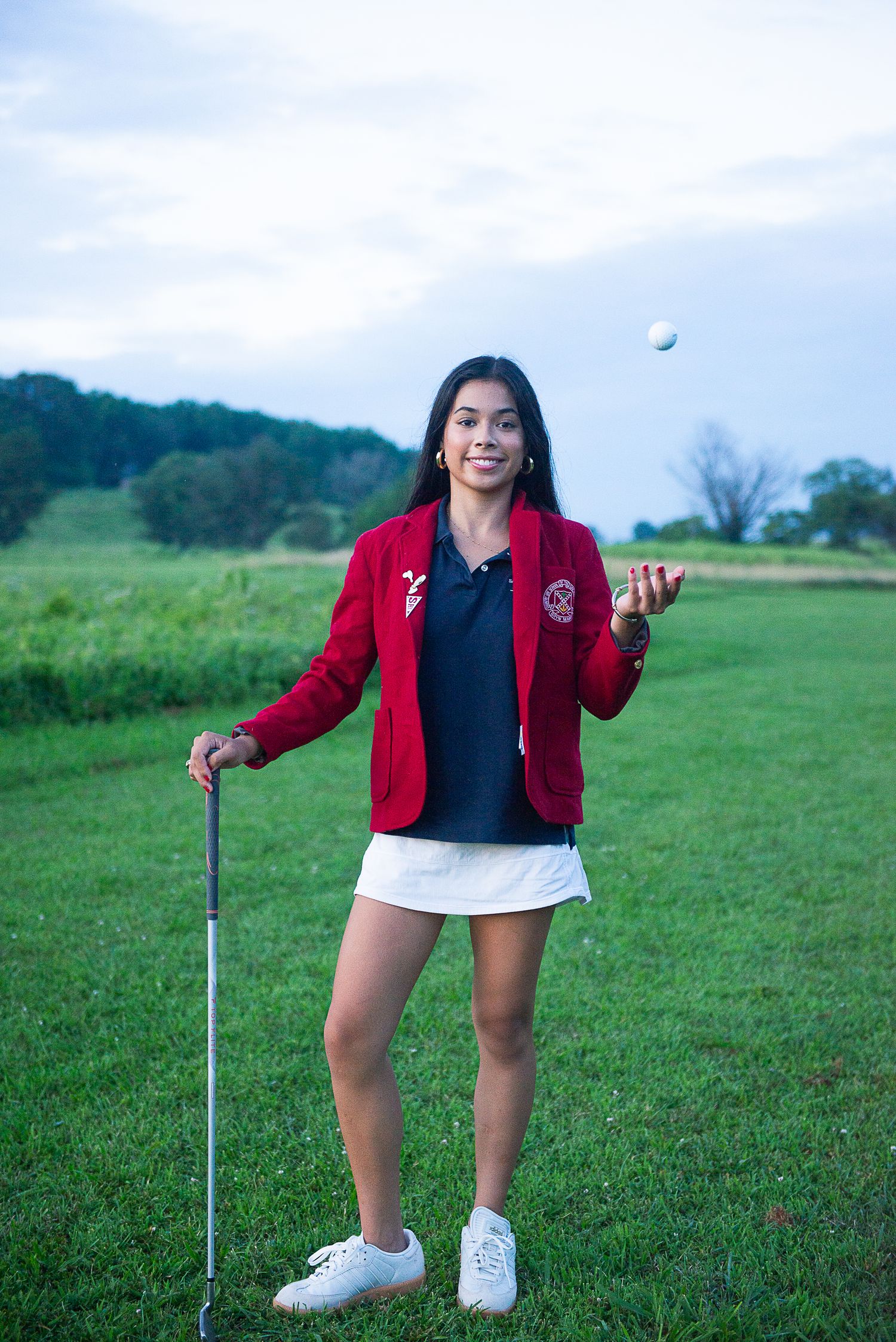 A golfer in a red blazer and white skirt playfully tosses a ball on a scenic golf course.