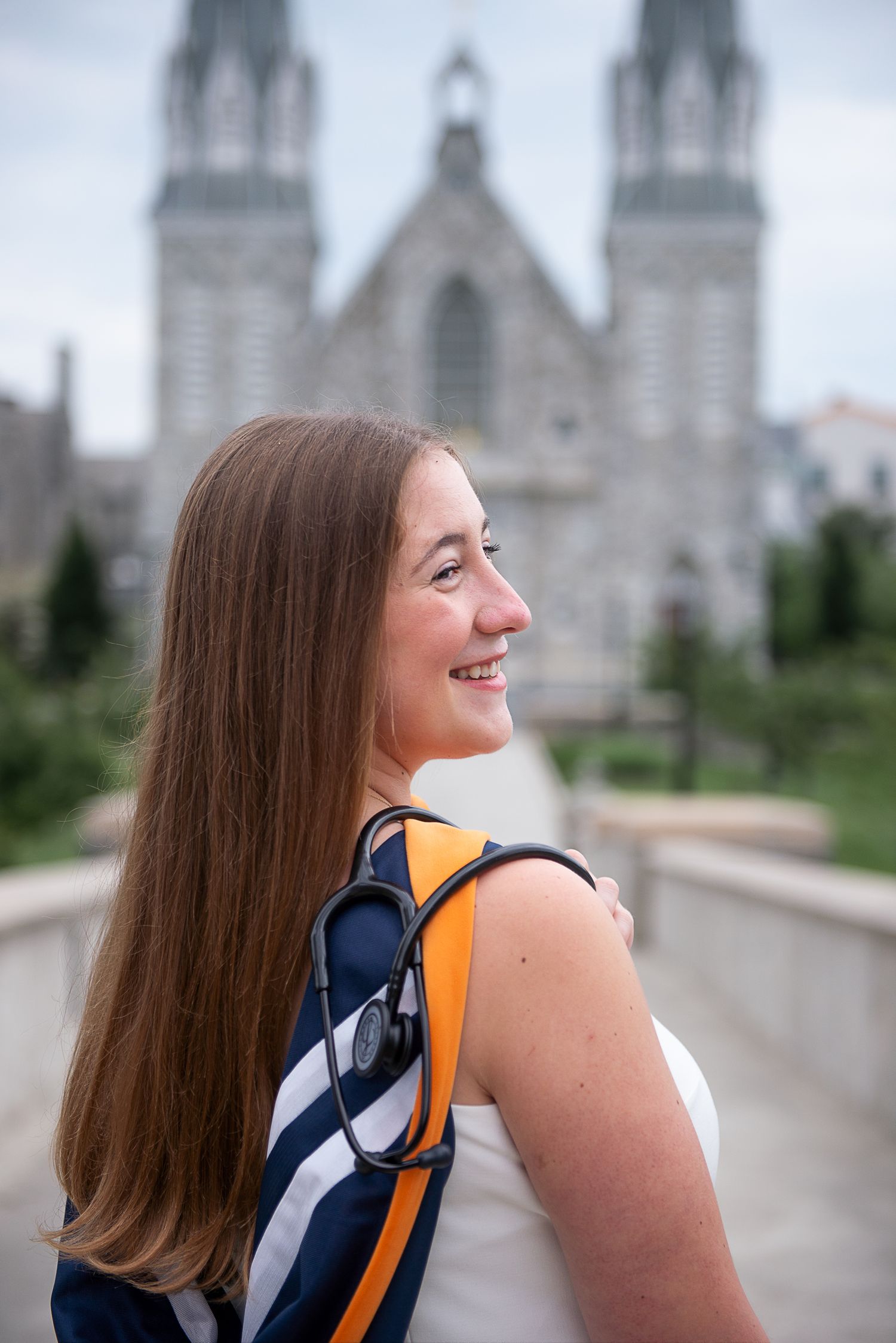 Graduate wearing sash looks back over shoulder with St Thomas church  in background on Villanova's campus.