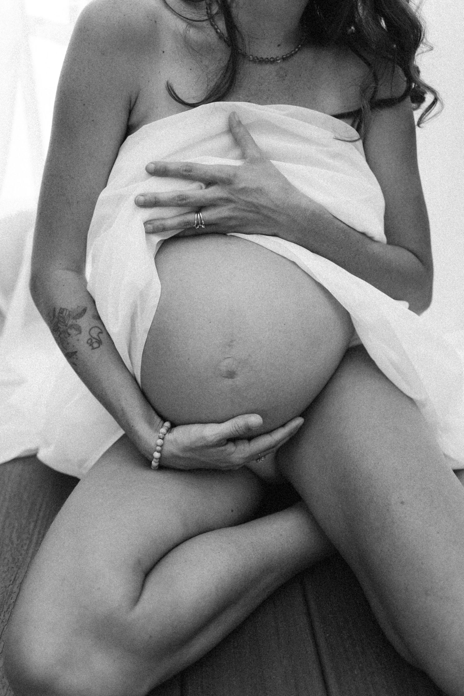 Black and white artistic maternity photo showing a pregnant belly wrapped in white fabric with hands gently cradling the bump.