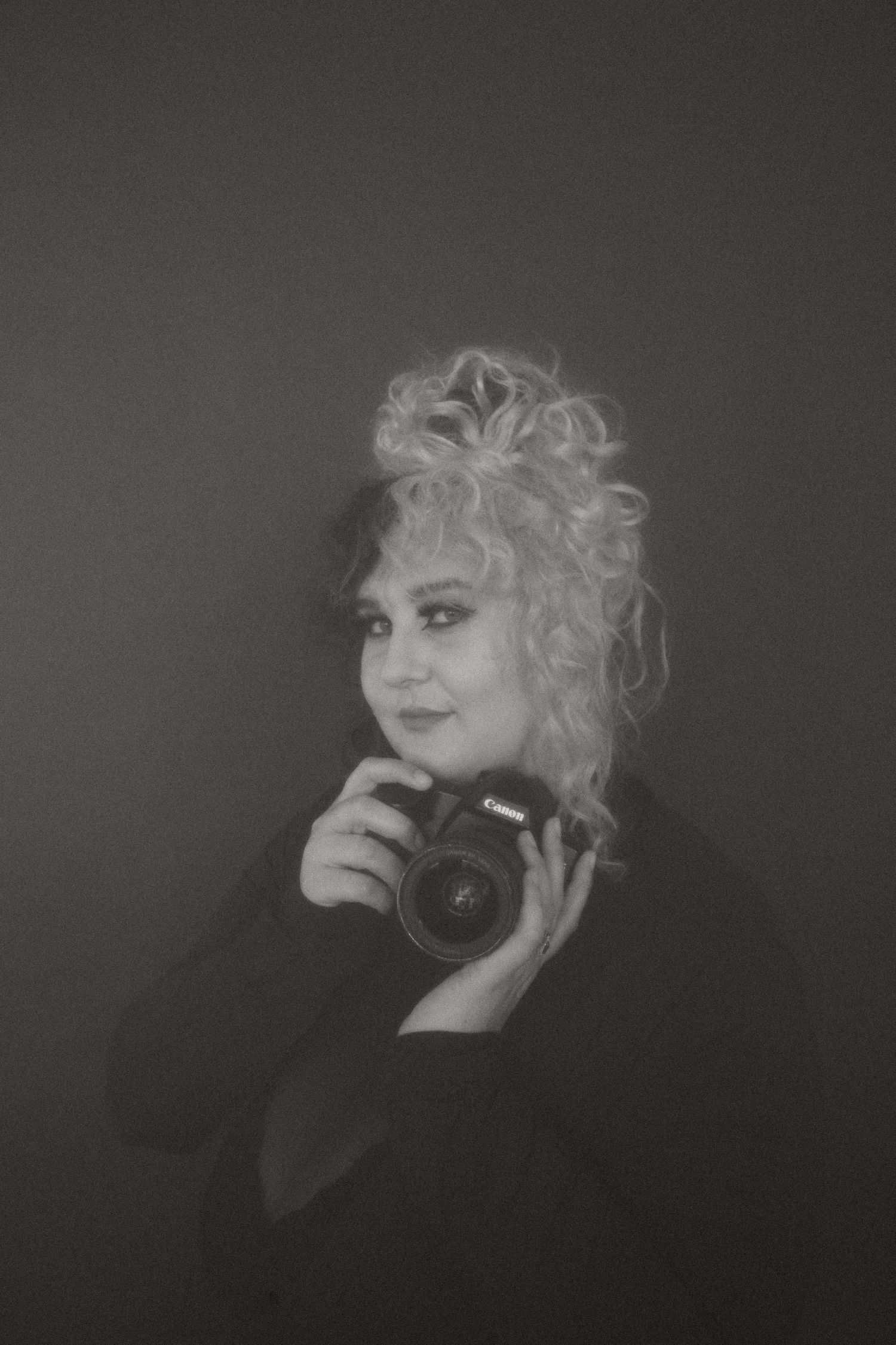 Black and white portrait photo of a photographer holding a camera in dark studio lighting with curled hairstyle.