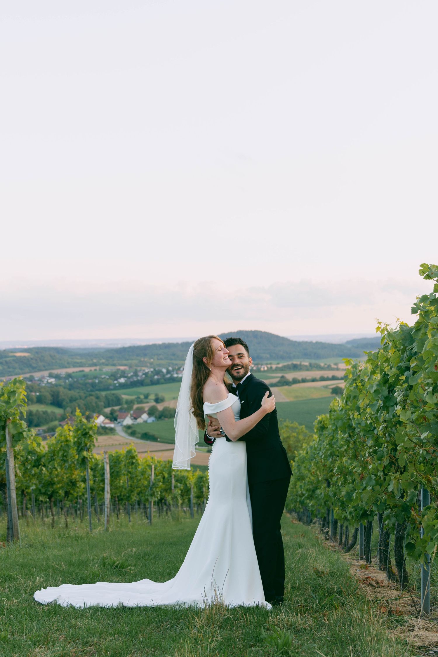 Newlyweds share romantic moments in a scenic hillside vineyard at sunset surrounded by rows of grapevines.