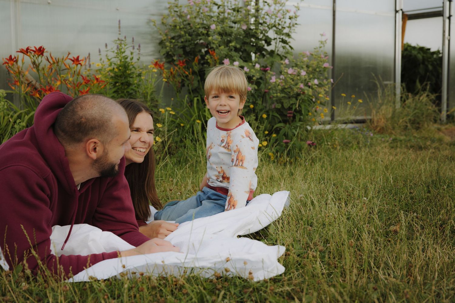 Family relaxing together on blanket in grassy yard with blooming flowers and greenery in background.