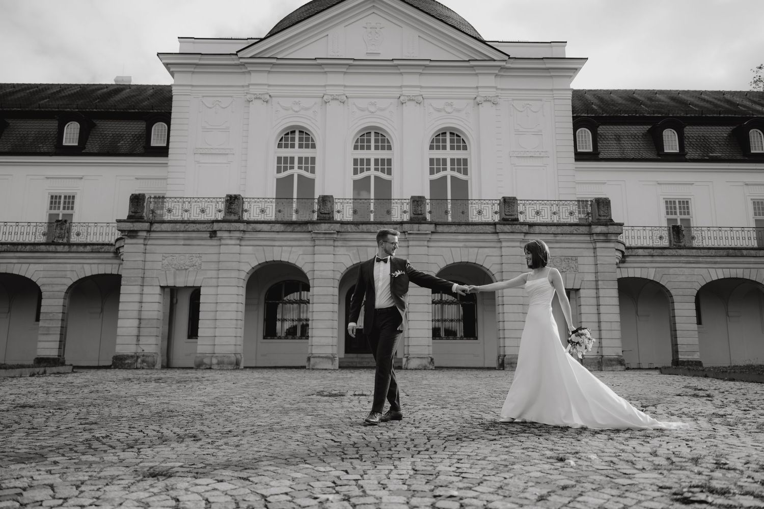 A couple posing in formal attire on cobblestone ground in front of a grand historic building.