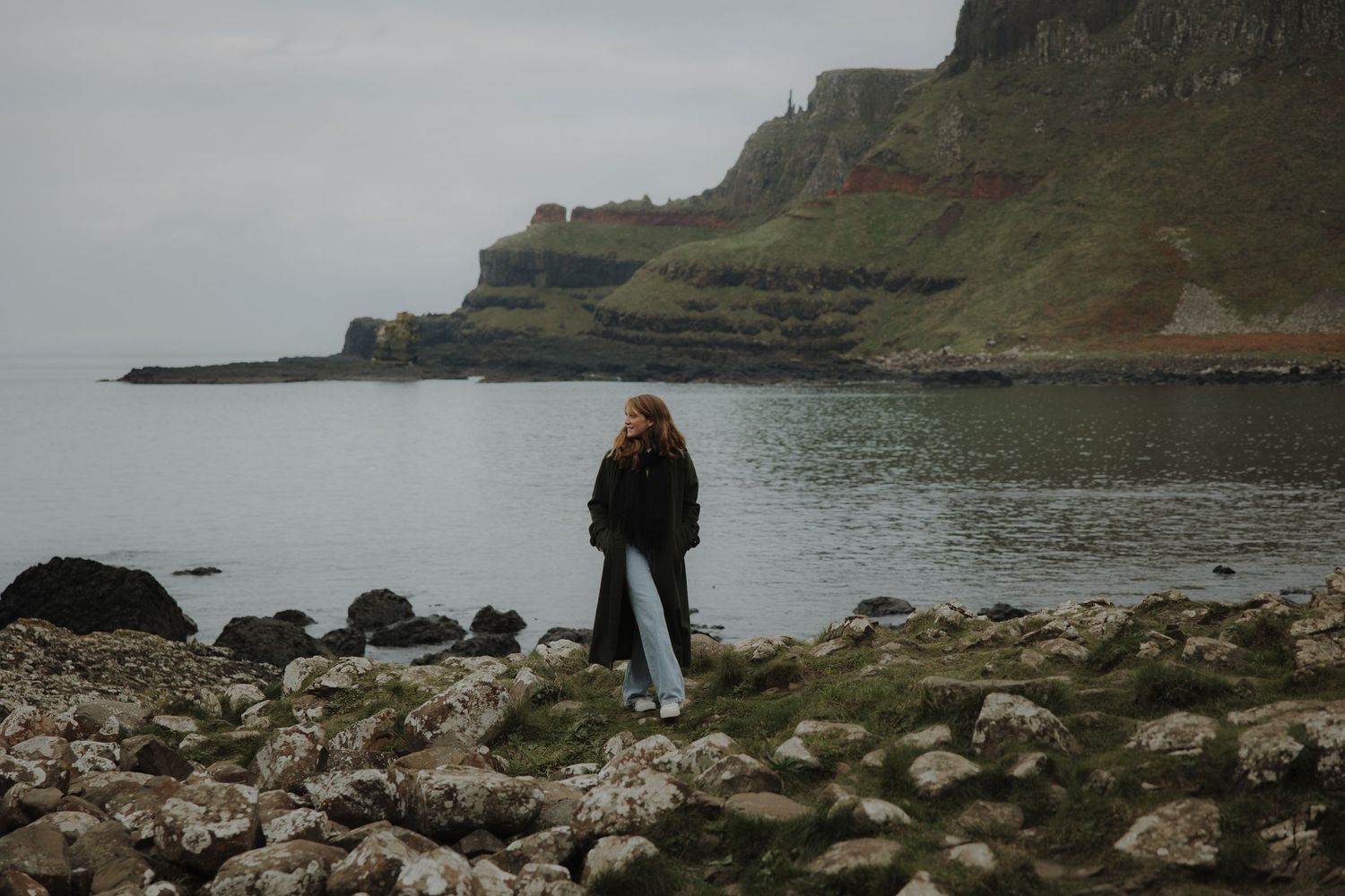 A person stands among rocky shoreline with dramatic coastal cliffs and moody gray waters in the background.