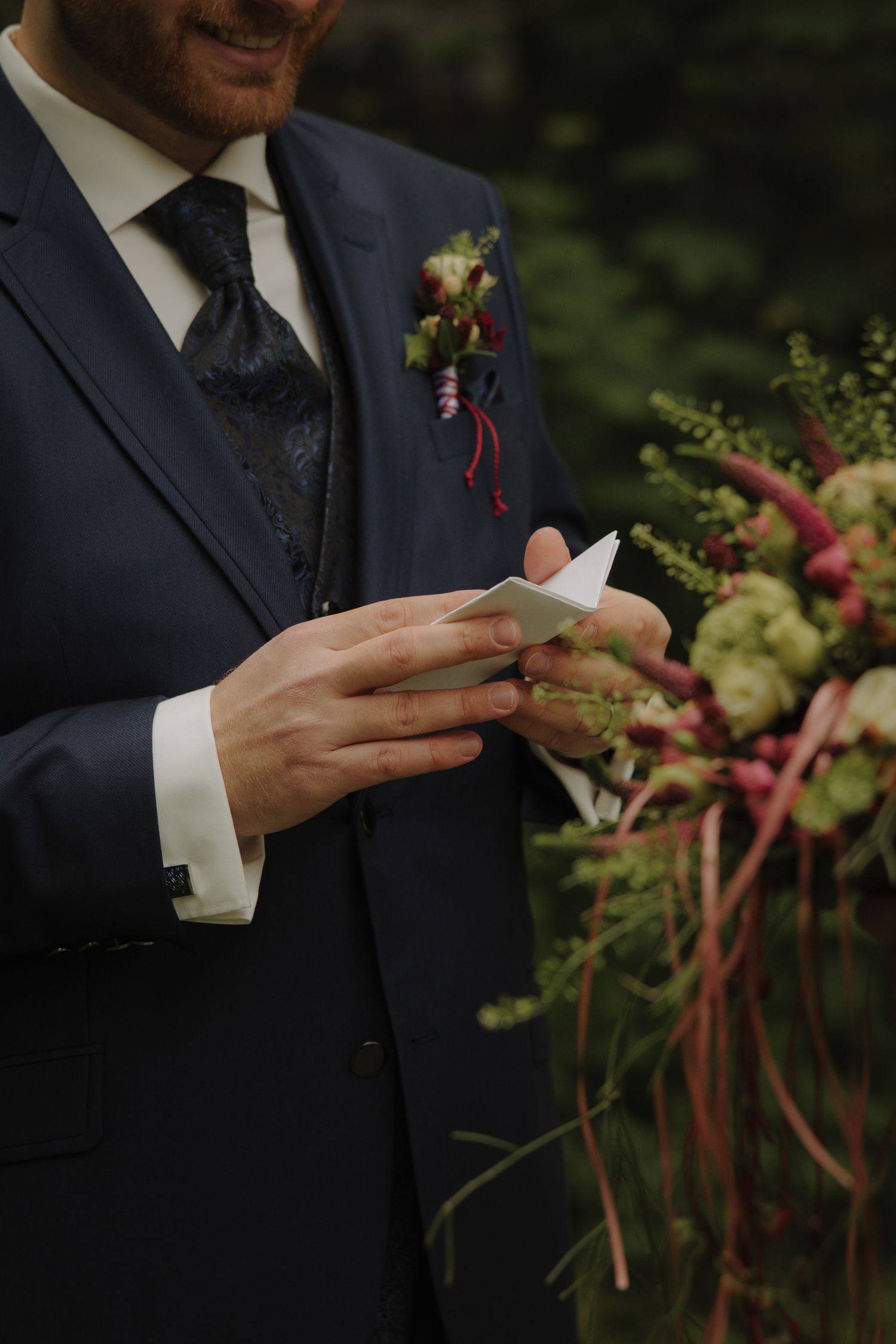 Groom in navy suit holding wedding vows note, with boutonniere and floral arrangement nearby.