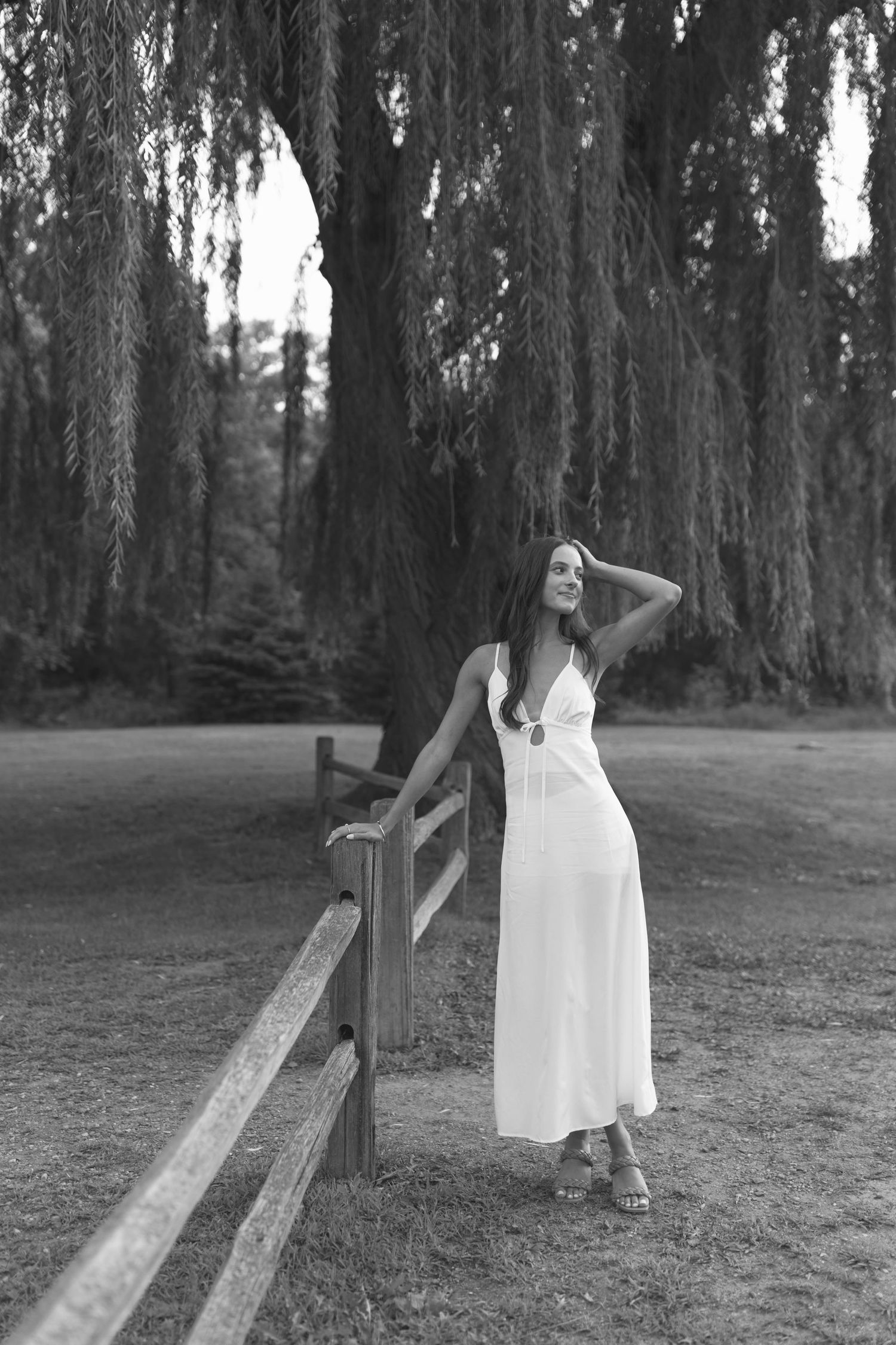 A person in a white dress stands near a tree in a moody black and white forest setting.