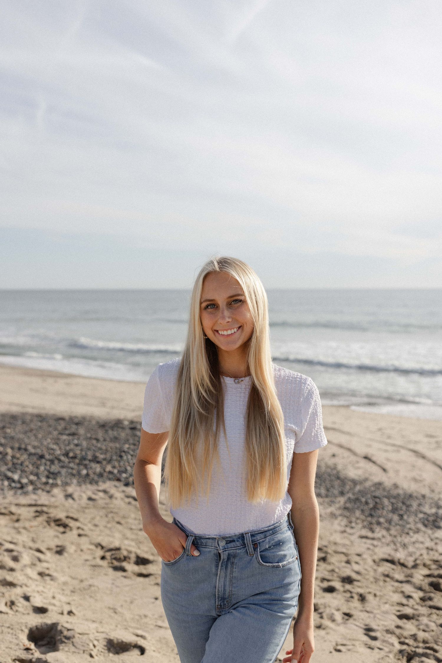 Woman in white t-shirt and jeans poses casually on rocky beach with ocean waves in background.