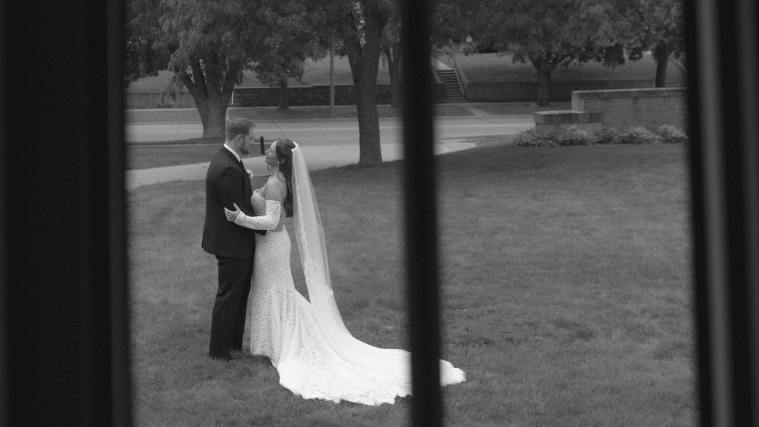 A romantic black and white wedding photo captured through a window frame in a garden setting.