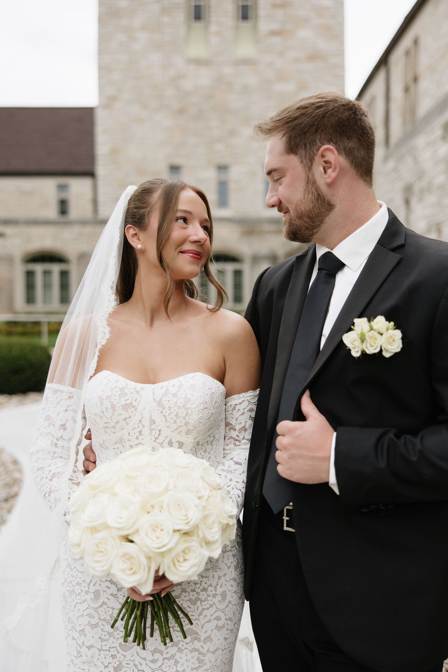 A bride in a lace wedding dress holds white roses while sharing a tender moment with her groom outside a stone building.