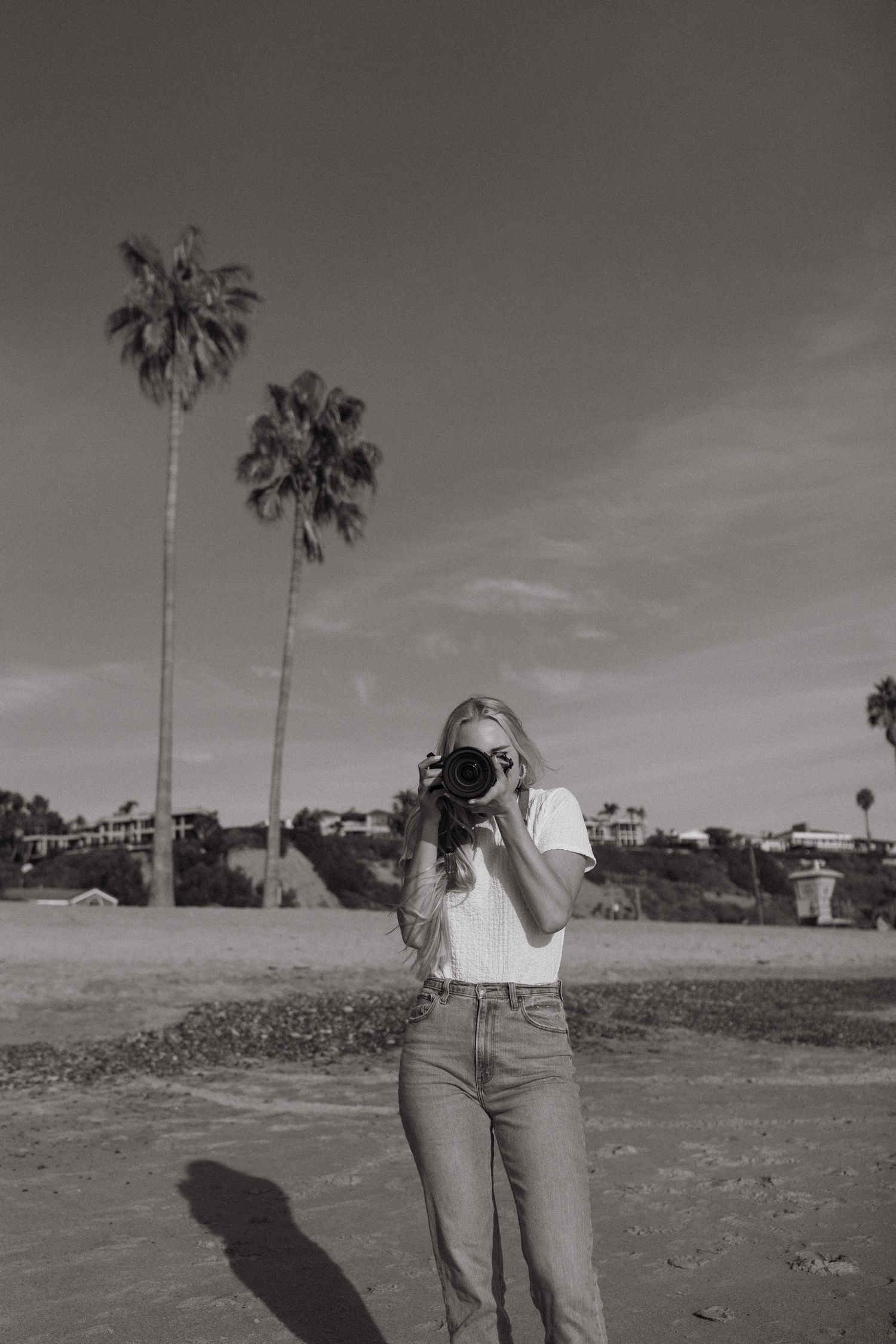 Black and white photo of a photographer with a camera against a palm tree backdrop in casual attire.