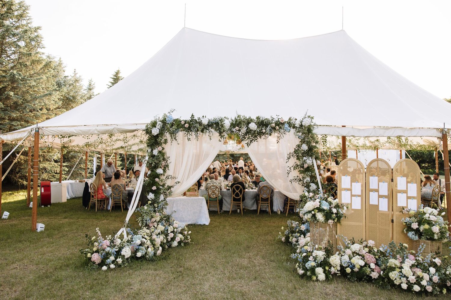 Elegant outdoor wedding tent decorated with floral arch, white drapes, and flower arrangements on green lawn.