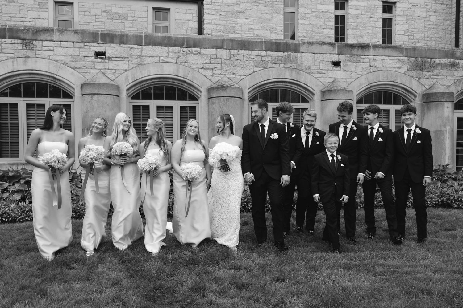 Black and white wedding party photo with bridesmaids in gowns and groomsmen in suits outside a stone building.