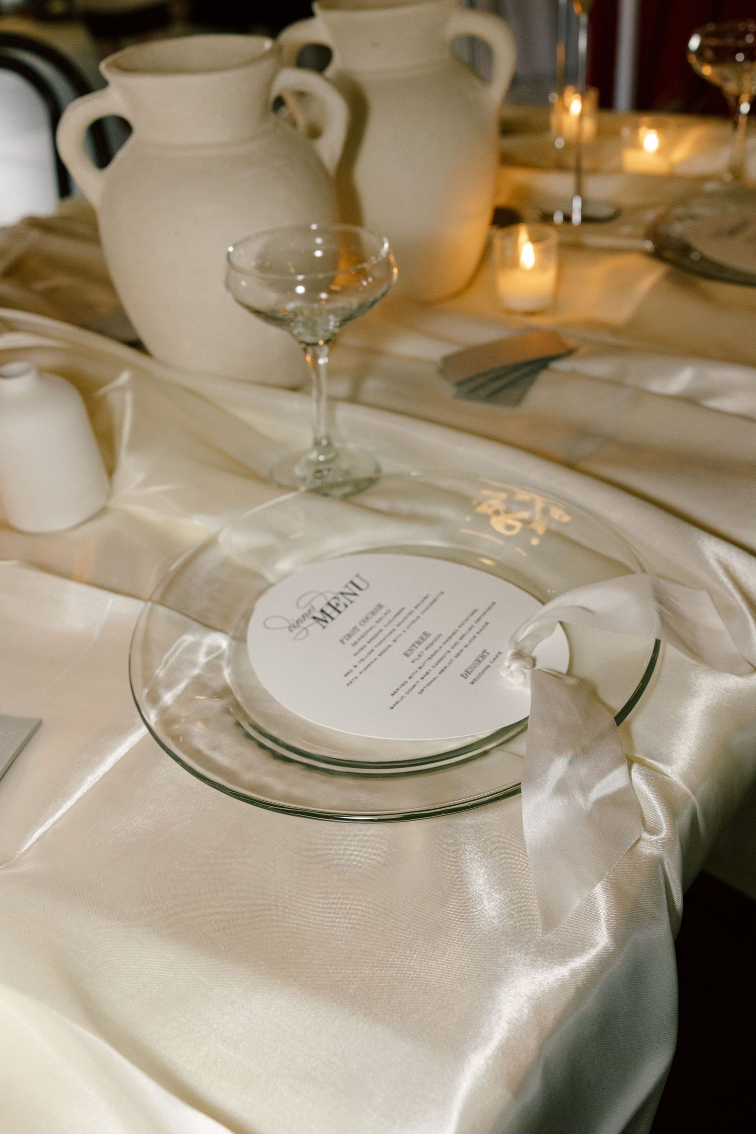 Elegant wedding place setting with menu card, wine glass and white ceramic pitchers.