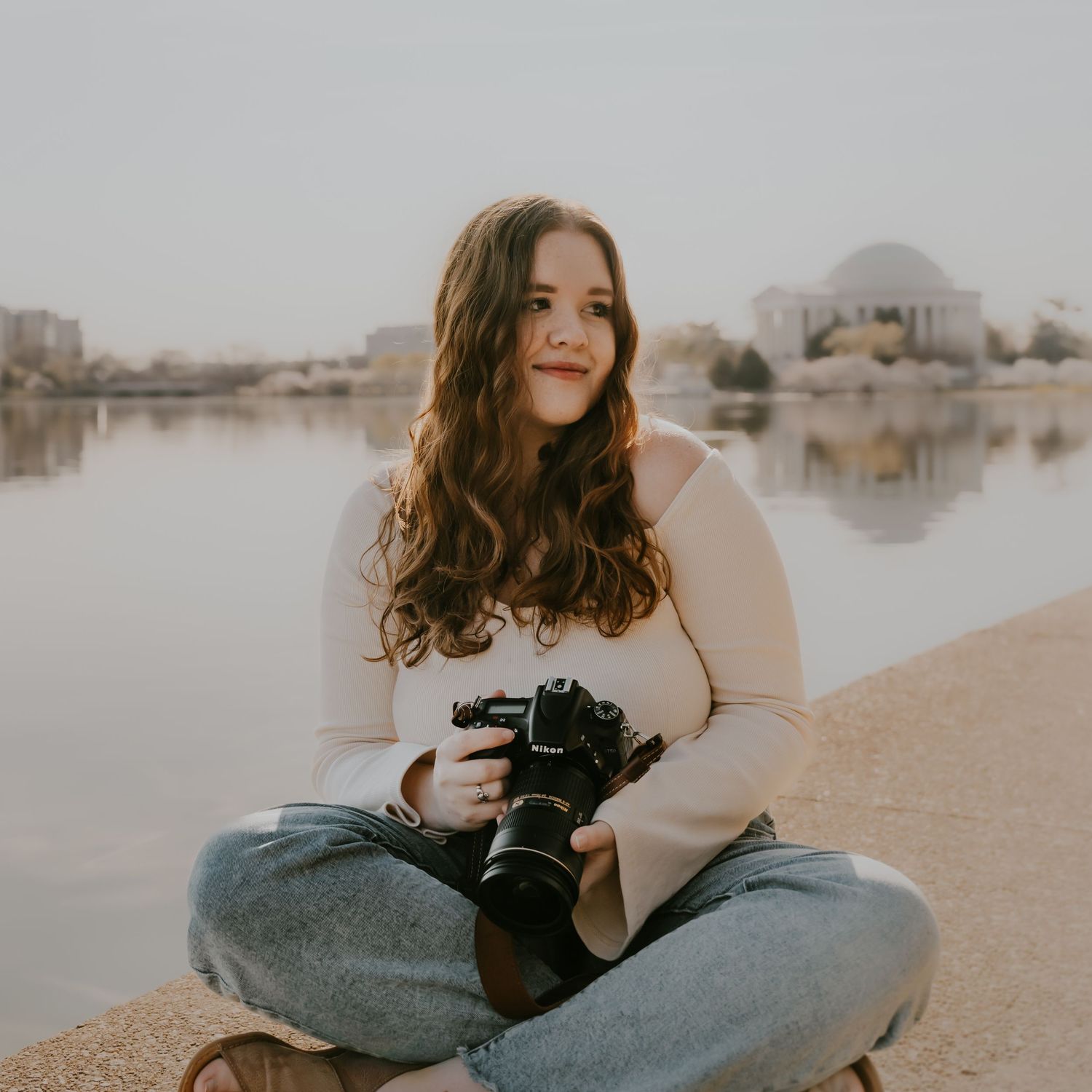 Photographer with long wavy hair sits cross-legged by a lake holding a professional camera at sunset.