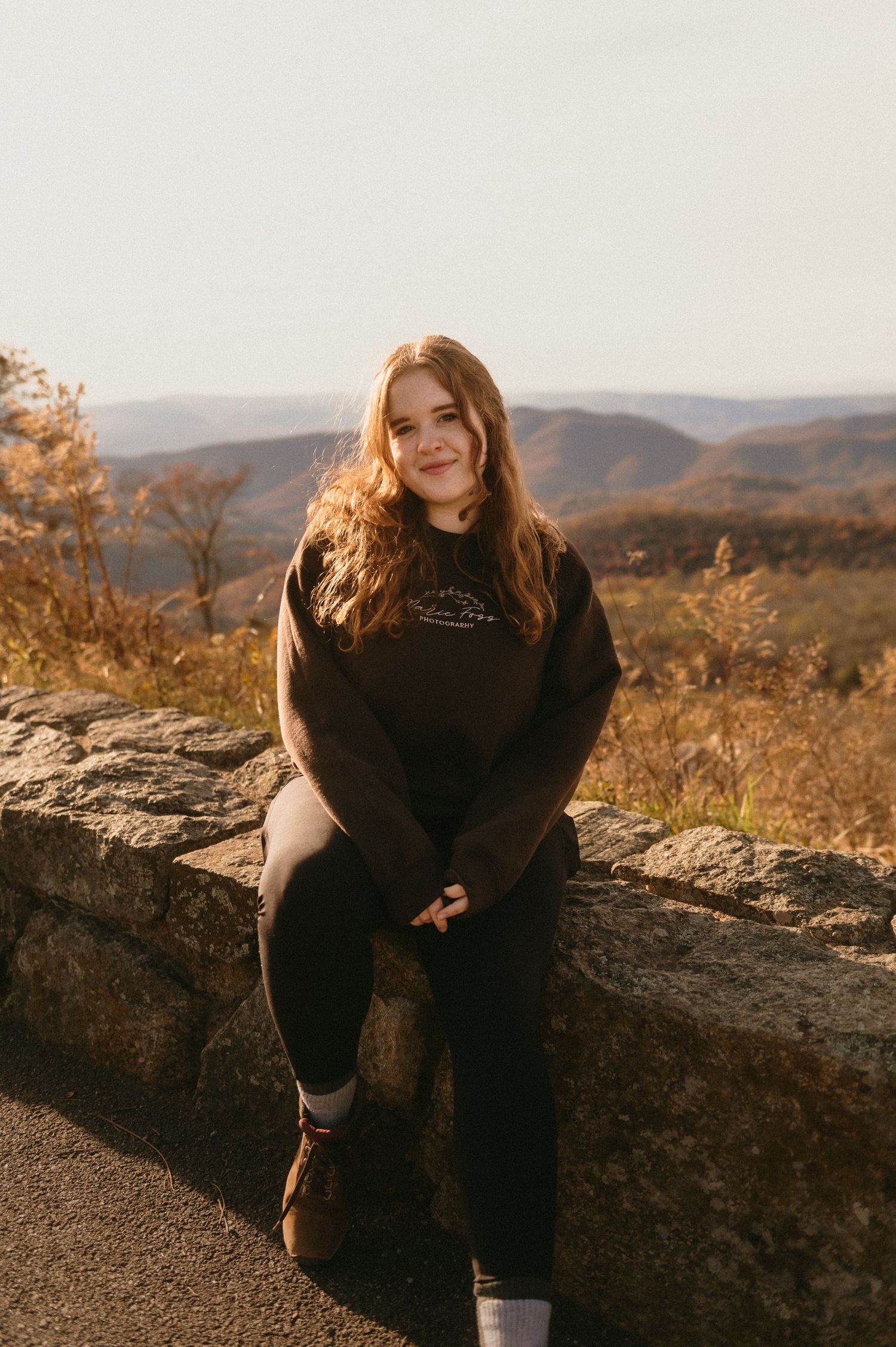 A person in black sits on a stone wall overlooking an autumn mountain landscape during golden hour.