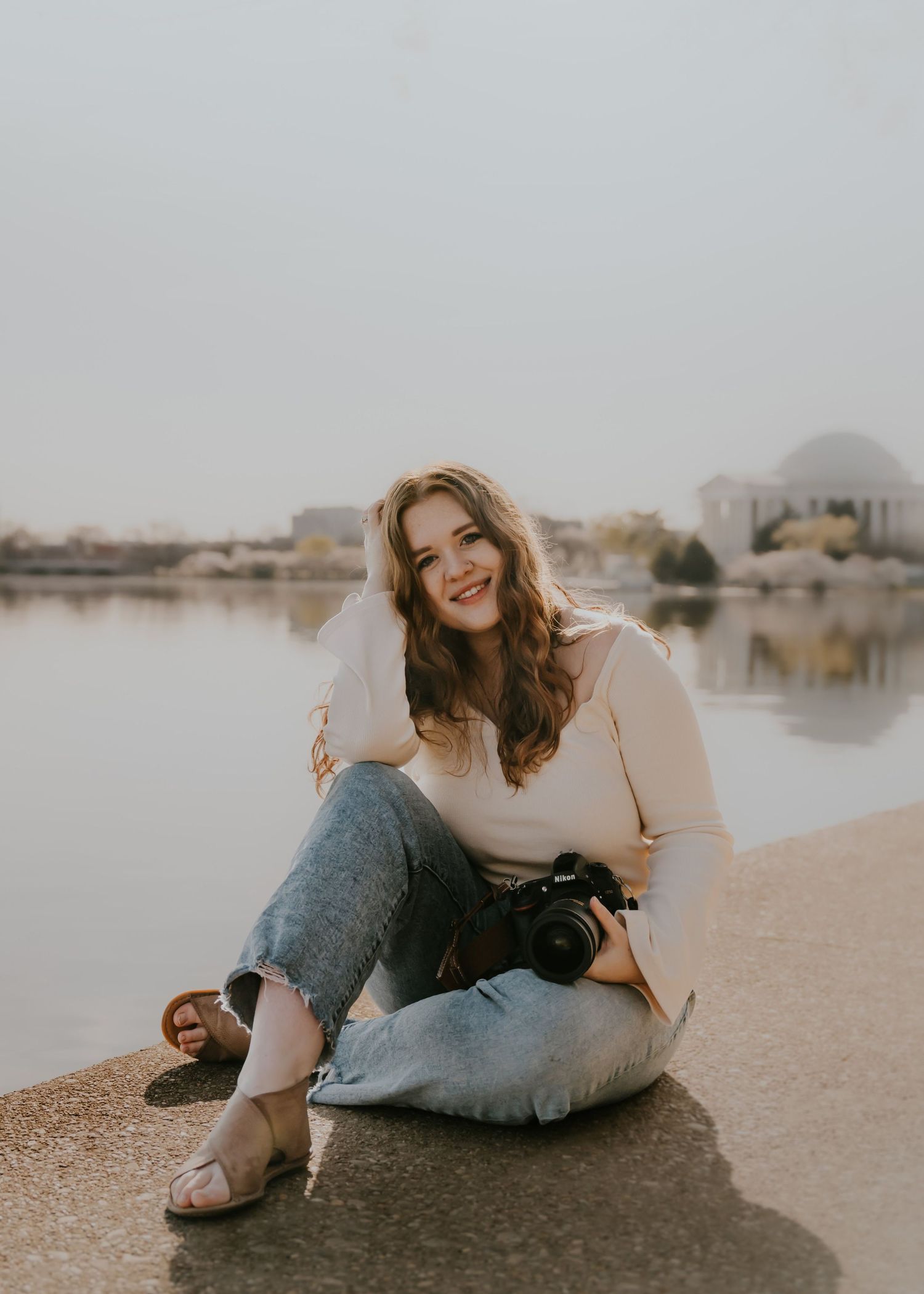 Person in jeans and cream sweater relaxes on sandy beach at sunset with water view in background.