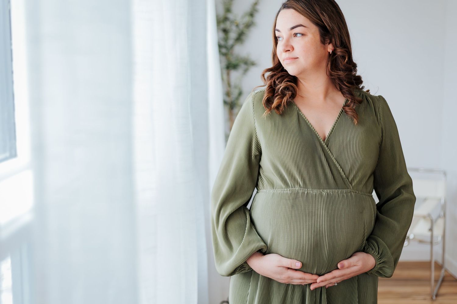 Pregnant woman in green dress cradling baby bump, standing by window in bright room. Pregnancy dresses for photoshoot. Maternity photography