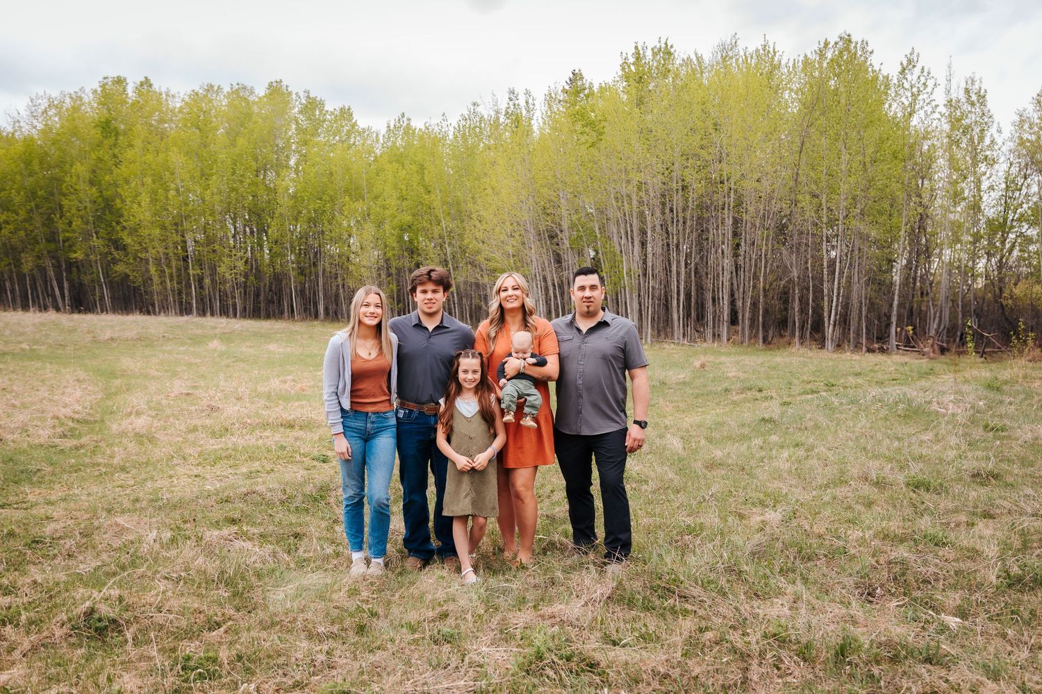 Extended family group of six smiling together in green meadow surrounded by spring trees.
