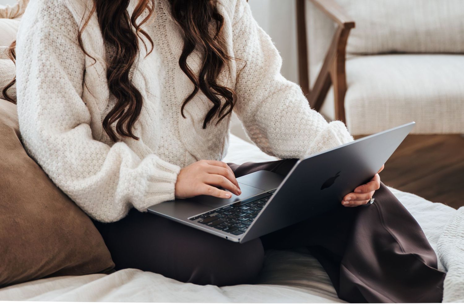 Woman in cozy white sweater sitting cross-legged on bed, typing on laptop computer in bright, comfortable home setting. business photos.