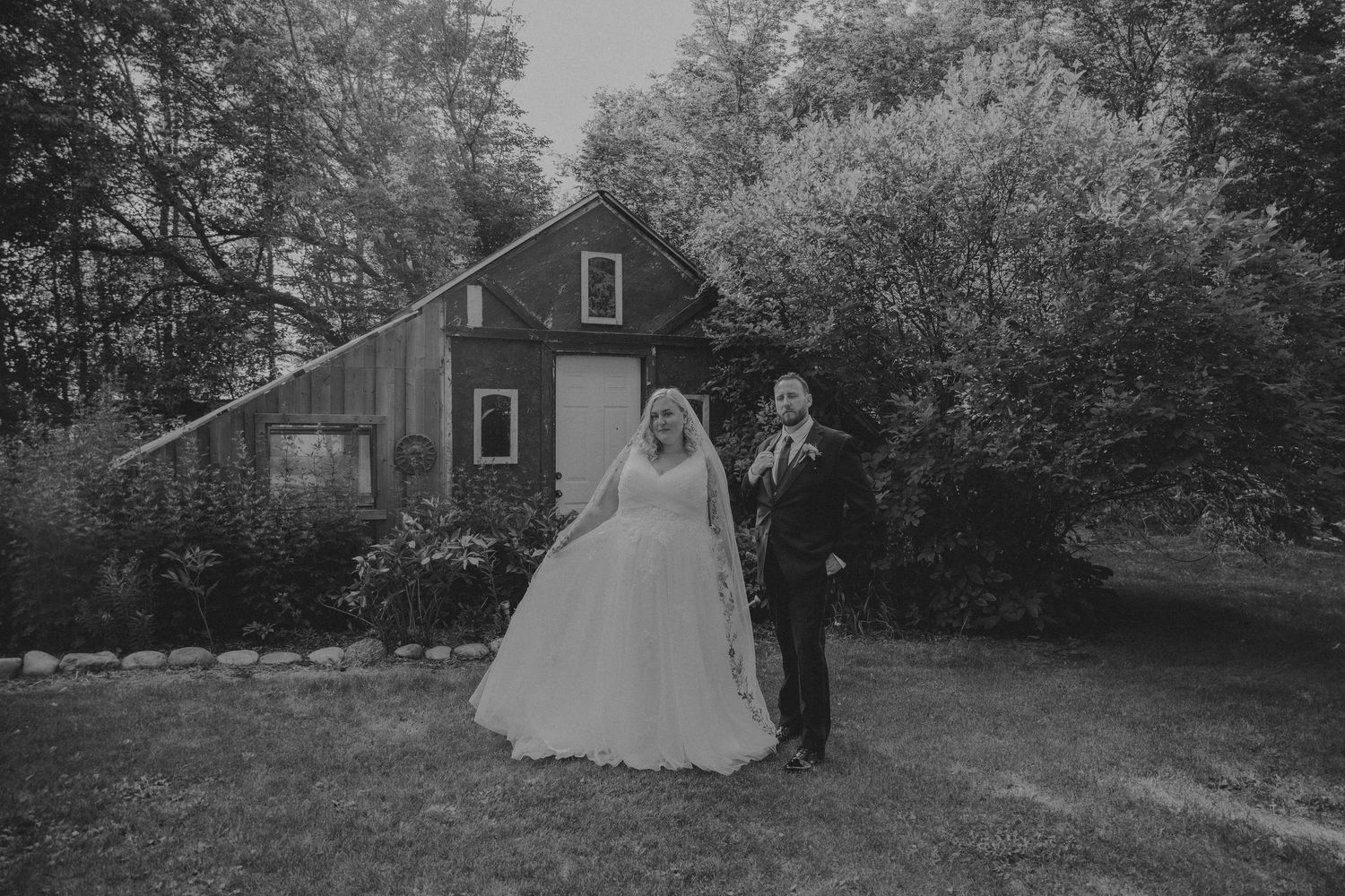 Black and white photo of a bride and groom holding hands in front of a rustic barn surrounded by lush trees.